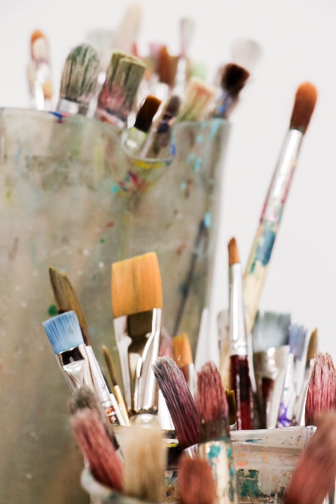 Close-up of various paintbrushes with colorful, paint-stained bristles in containers. Brushes vary in size and bristle shape, all standing upright, suggesting a busy creative environment. The background is softly blurred.