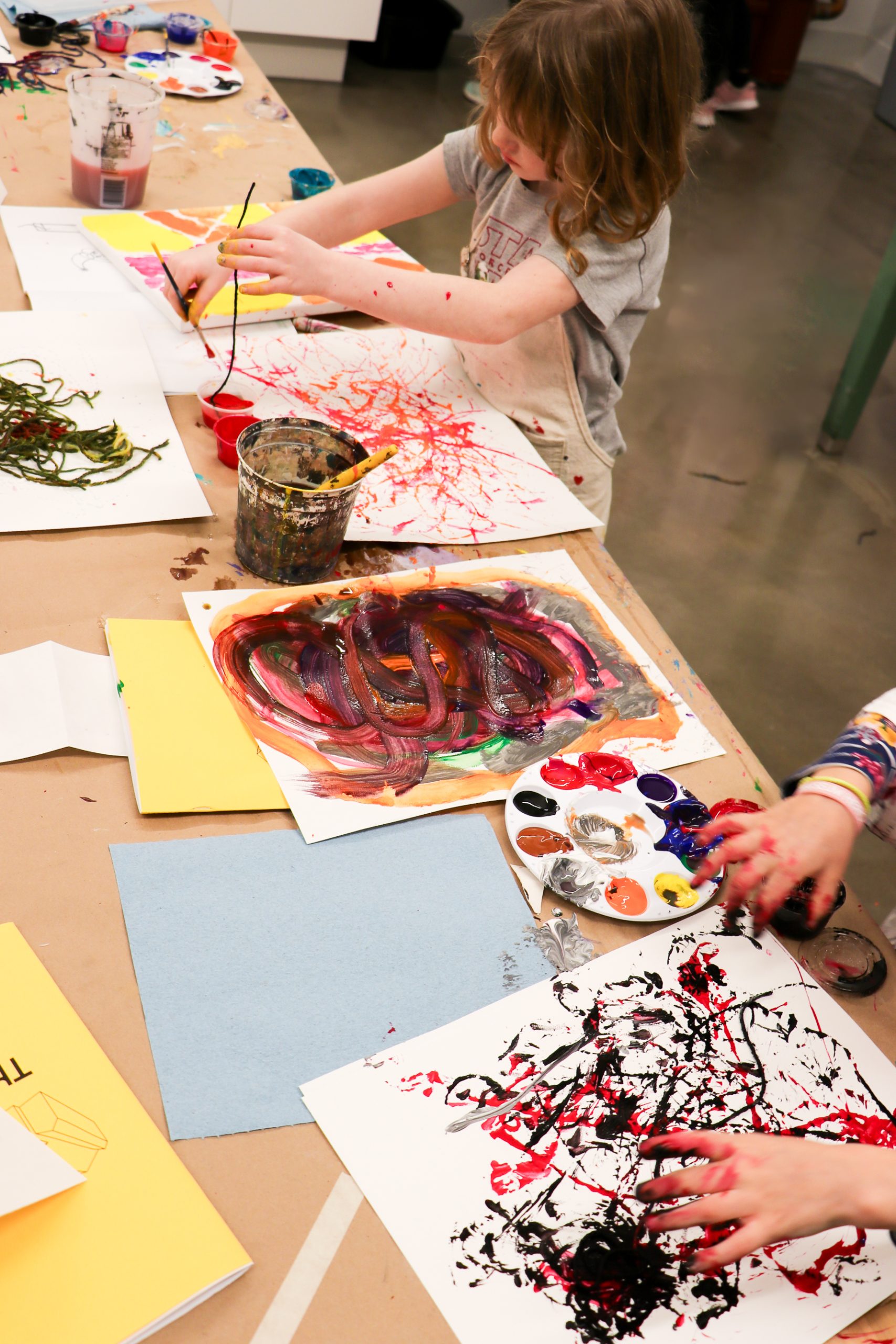 Two children engaged in painting activities at a table. They are working on colorful abstract paintings using brushes and paint. The scene is creative and vibrant, with art supplies scattered around.
