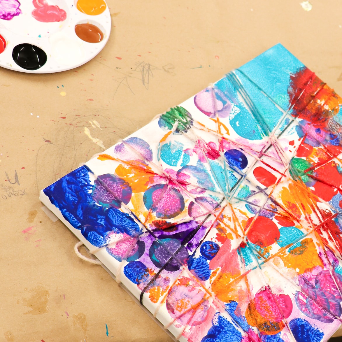 A vibrant, abstract painting with vivid colors in shades of blue, pink, orange, and purple on a canvas. Nearby, a palette with dabs of various paints rests on a beige surface. The artwork features overlapping circular and flower-like shapes.