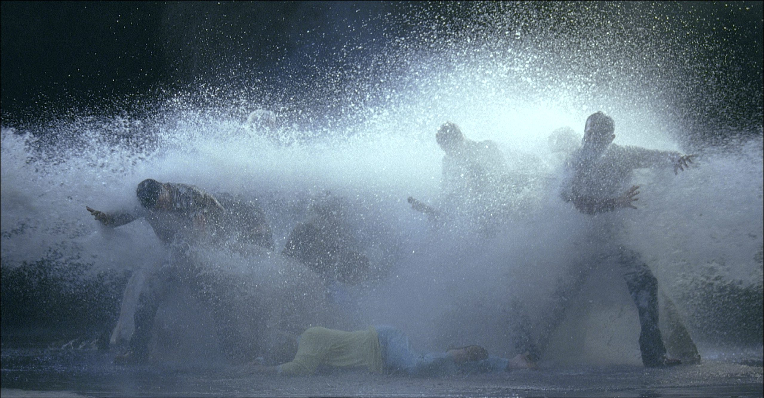 A group of people are dramatically illuminated as they are splashed with water. The scene is energetic and chaotic, with water droplets suspended in the air, capturing a moment of intense movement. One person is lying on the ground.