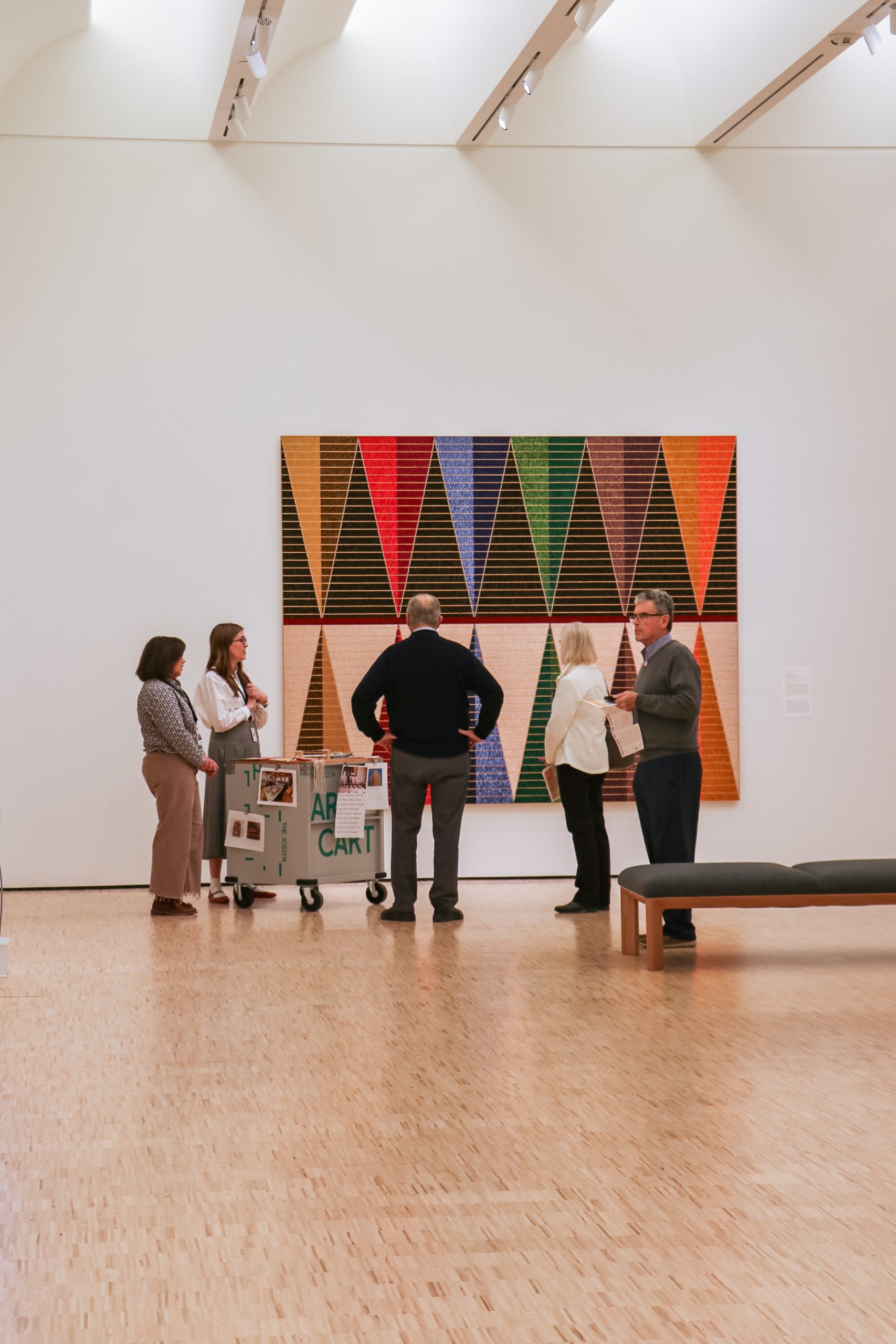 Five people are gathered in a gallery observing a large, colorful geometric artwork featuring triangular patterns. A mobile art cart is nearby. The gallery has a wooden floor and white walls with ceiling lights above.