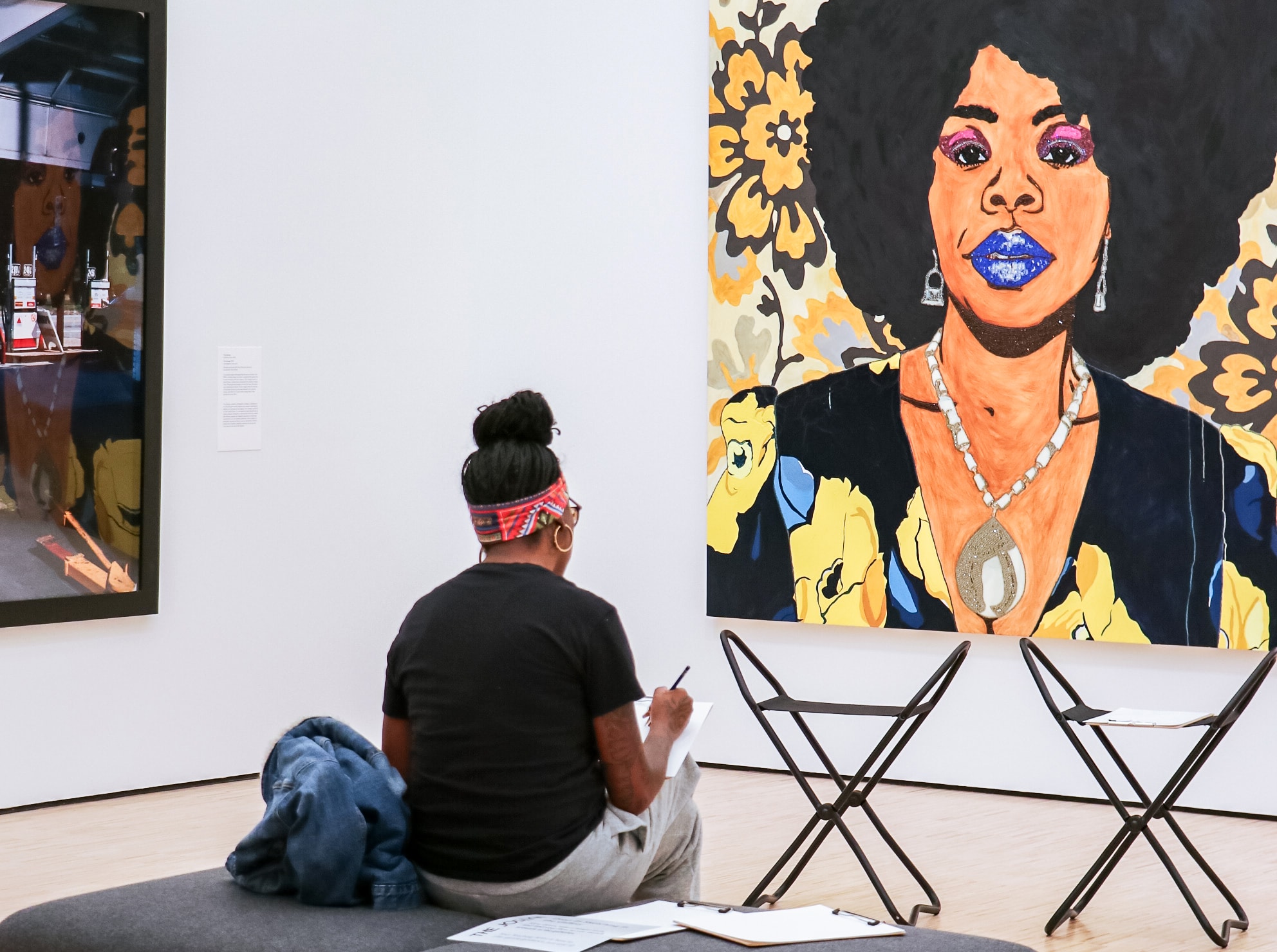 A person sits on a bench in an art gallery, sketching a large, colorful painting of a woman with an afro. The vibrant artwork features bold florals and striking colors. Two foldable stools are nearby.