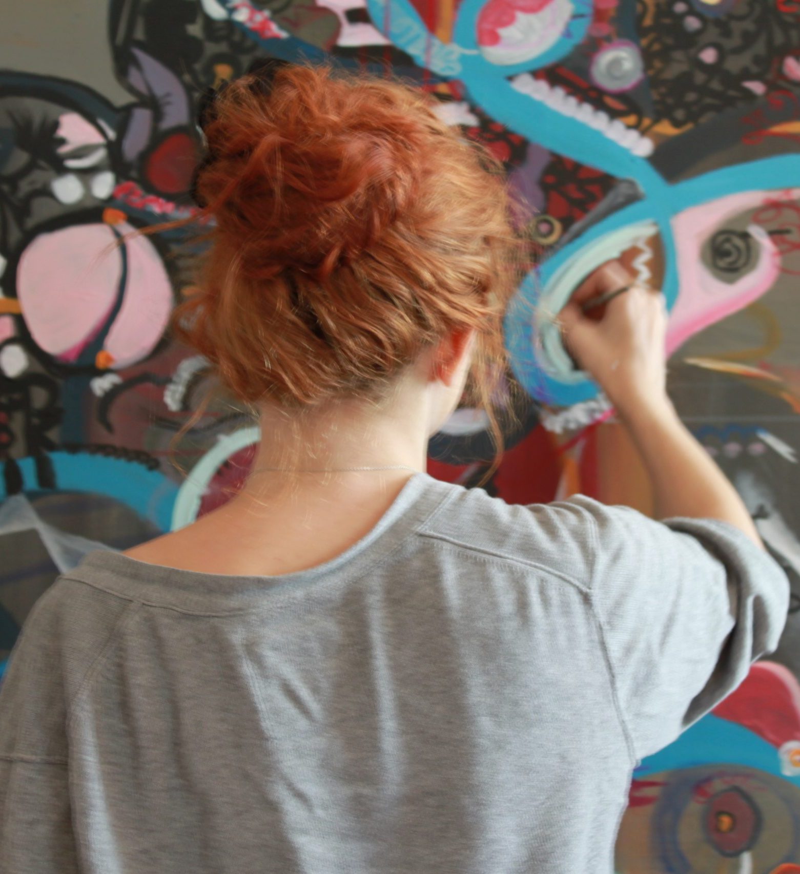 Person with curly, reddish hair painting a colorful abstract mural on a large canvas. The artwork features swirling patterns and vibrant colors. They are wearing a gray shirt and facing the mural.