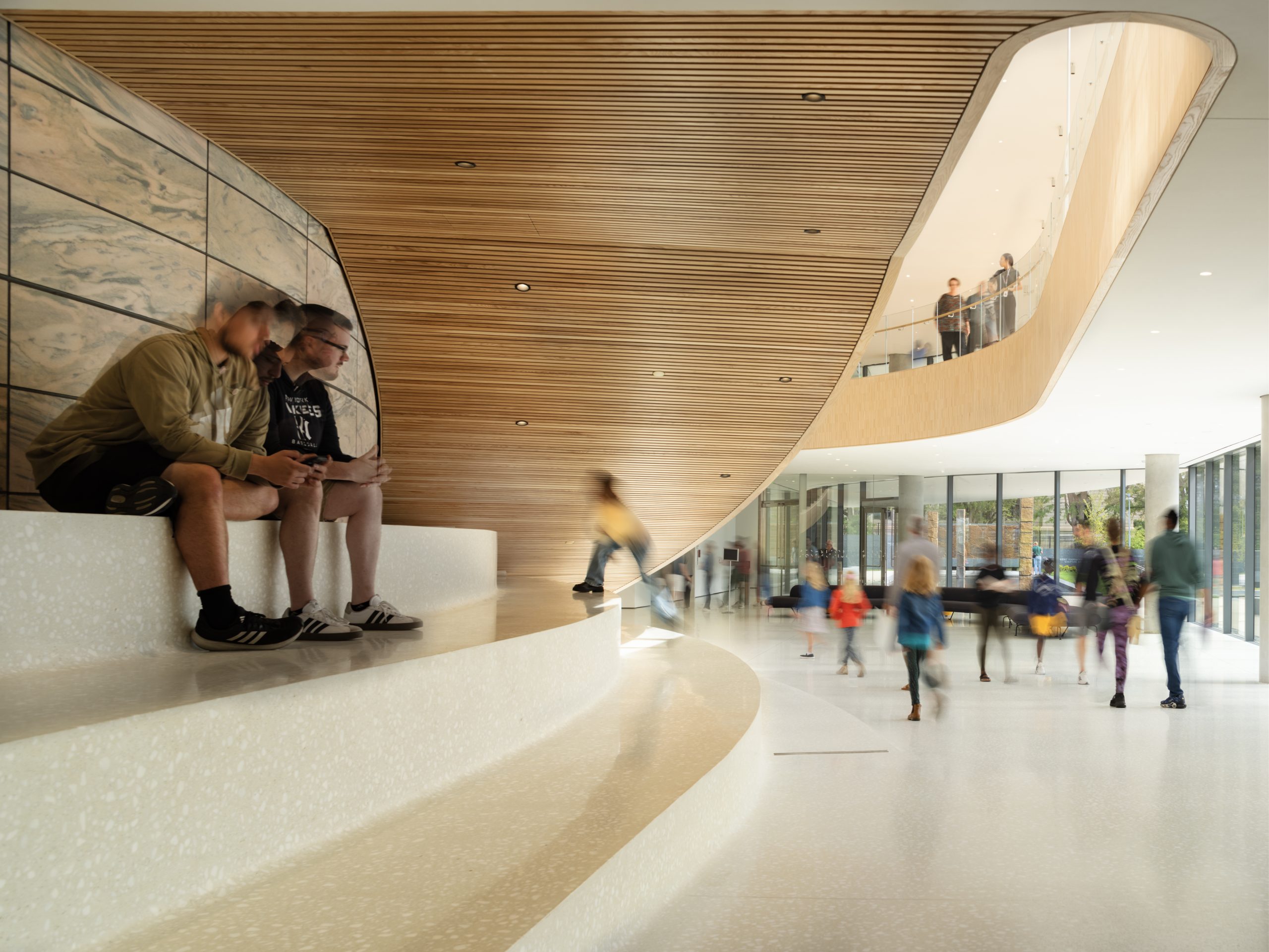 A modern interior with beige and wooden curved ceiling. Two people sit on large white steps using a smartphone. The background features a mezzanine, glass walls, and several blurred figures walking, creating a lively atmosphere.