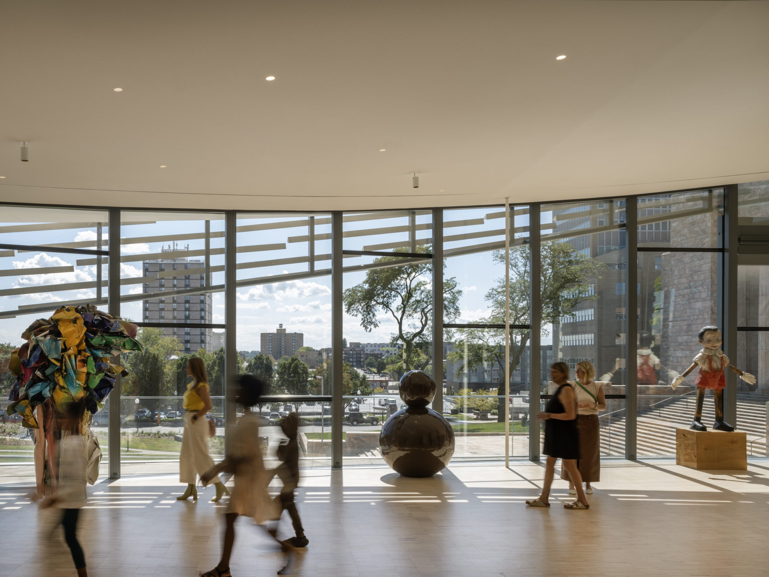 People walking in a spacious, modern gallery with large floor-to-ceiling windows, allowing natural light to illuminate sculptures and art pieces. Urban buildings and trees are visible through the windows, providing an outdoor view.