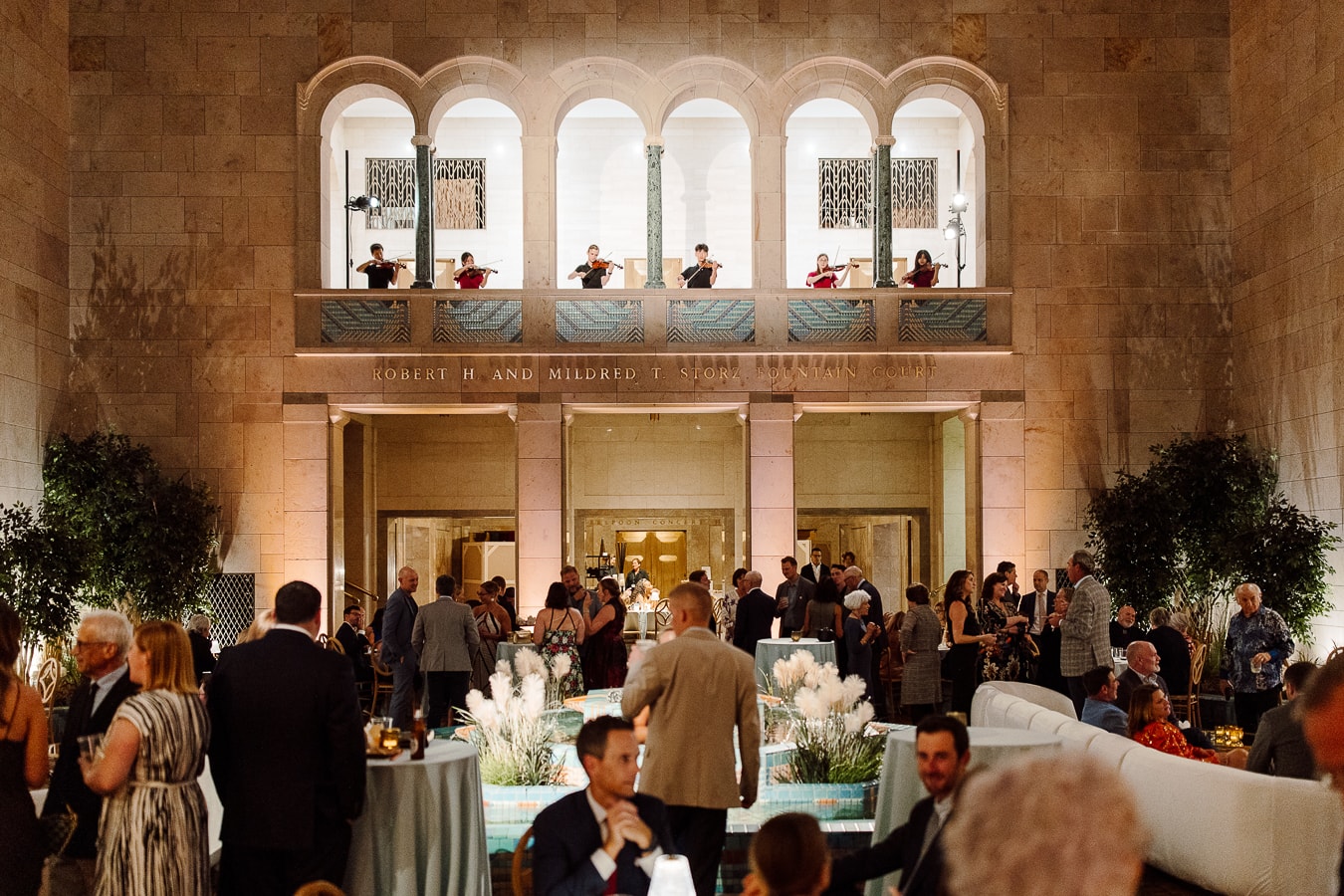 A large gathering in a grand hall with arched balconies and classical architecture. People are mingling and seated at tables, while musicians play on a balcony above. The room is warmly lit, enhancing the elegant ambiance.