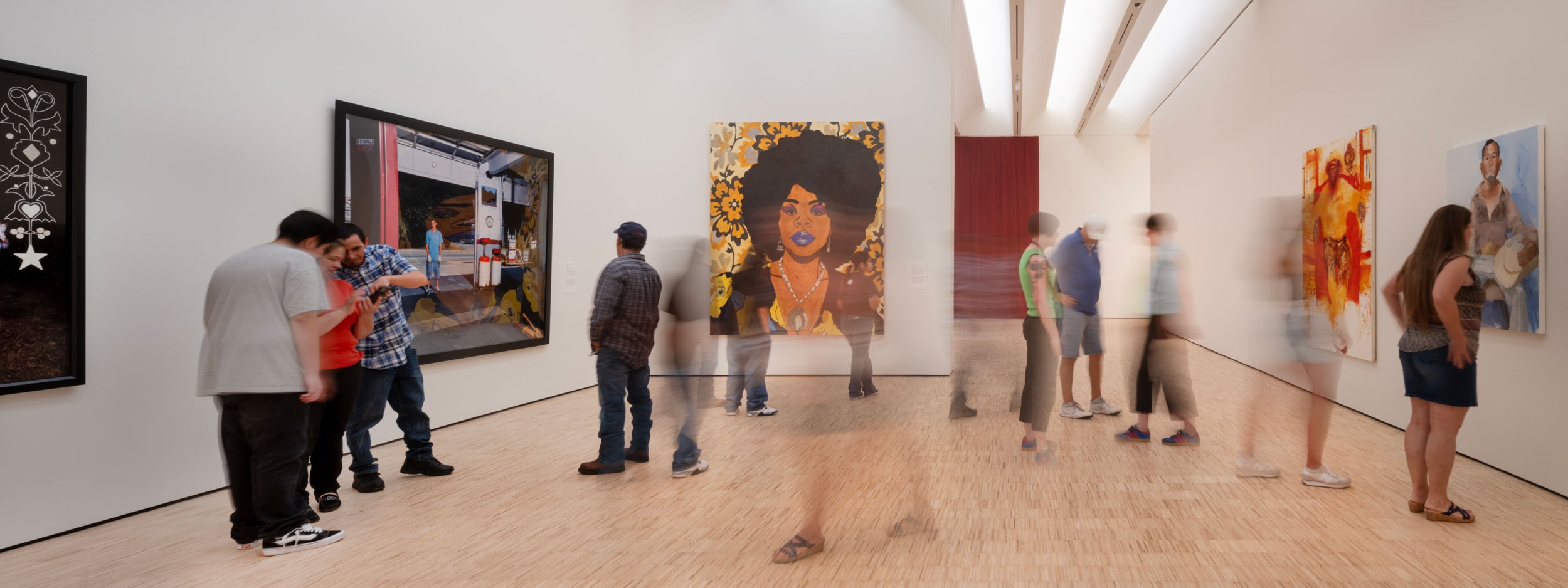 Visitors view art in a modern gallery. The walls display colorful paintings, including a prominent portrait of a woman with an afro. Some people appear blurred, suggesting movement. The floor is light wood, complementing the bright, spacious environment.