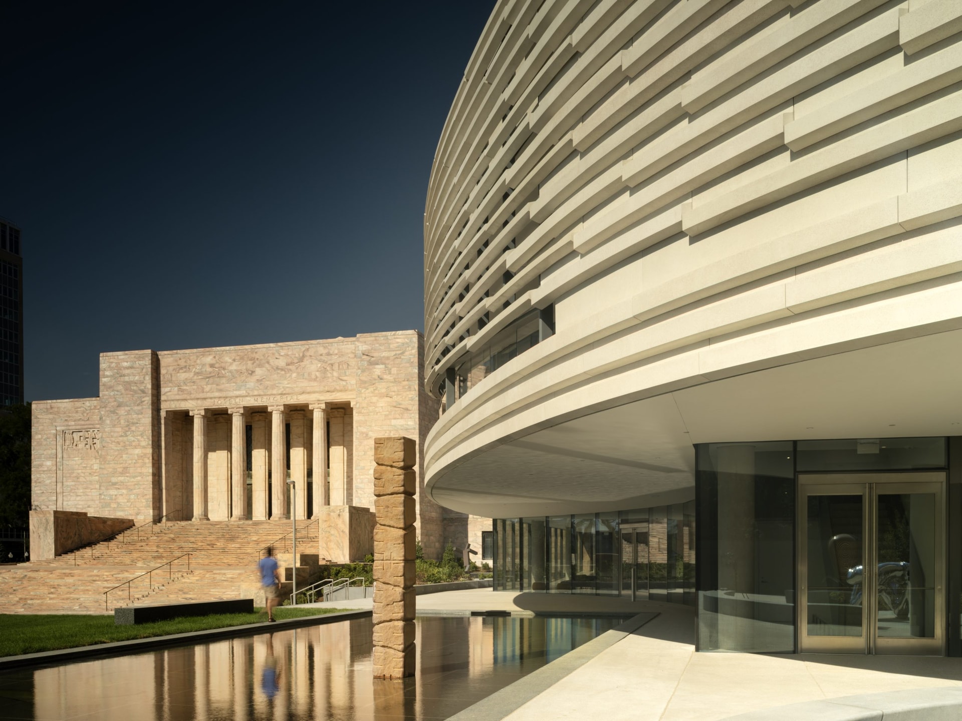 Modern curved building with reflective glass next to a historic structure with columns. A person walks nearby across a stone path over a reflective water feature, under a clear blue sky.