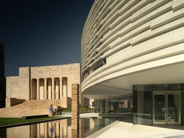 Modern curved building with reflective glass next to a historic structure with columns. A person walks nearby across a stone path over a reflective water feature, under a clear blue sky.