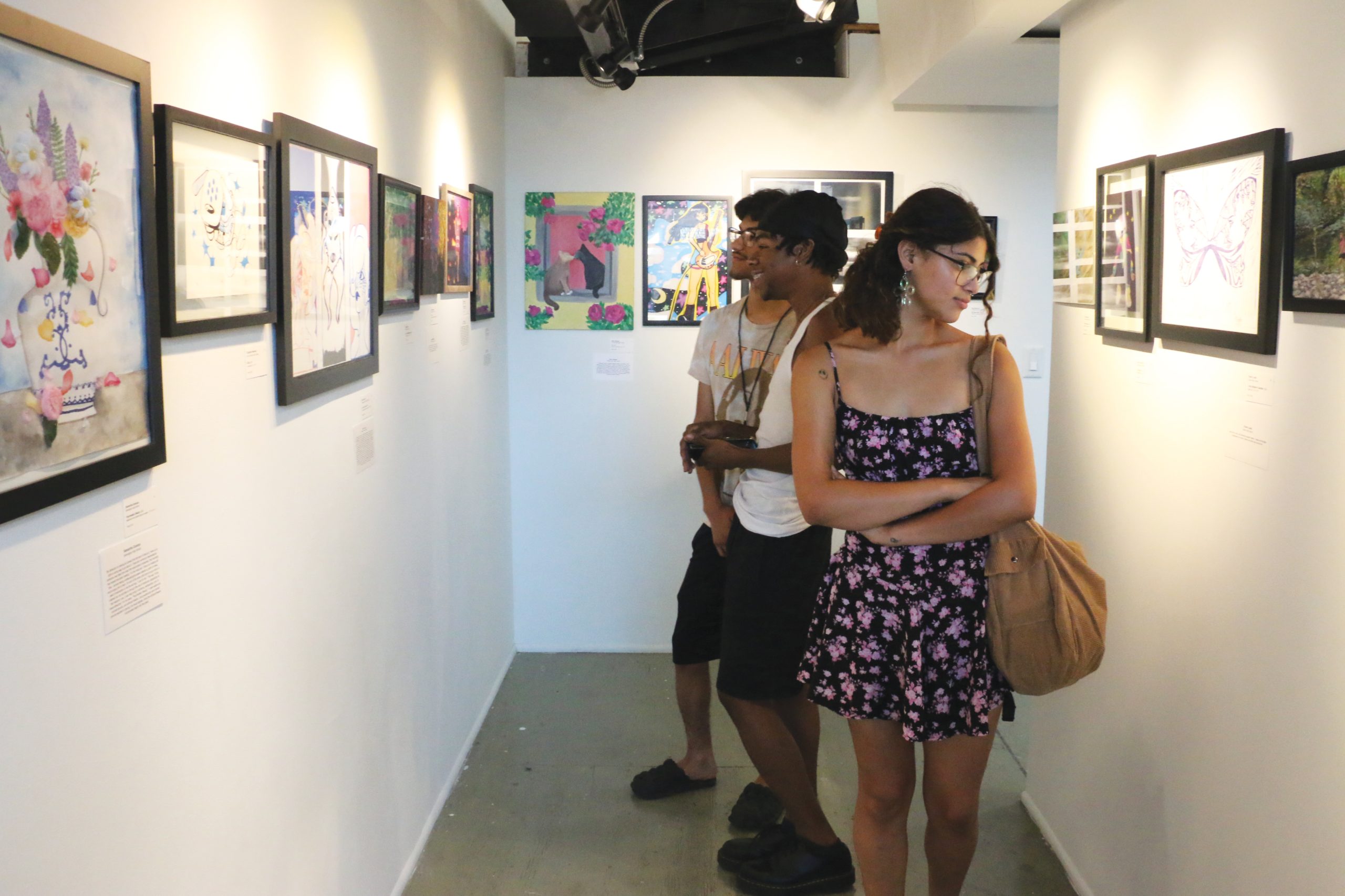 Three people in a gallery view colorful framed artworks on white walls. One person in a floral dress looks closely at a painting, while two others in casual attire engage in conversation. The setting is small and brightly lit.