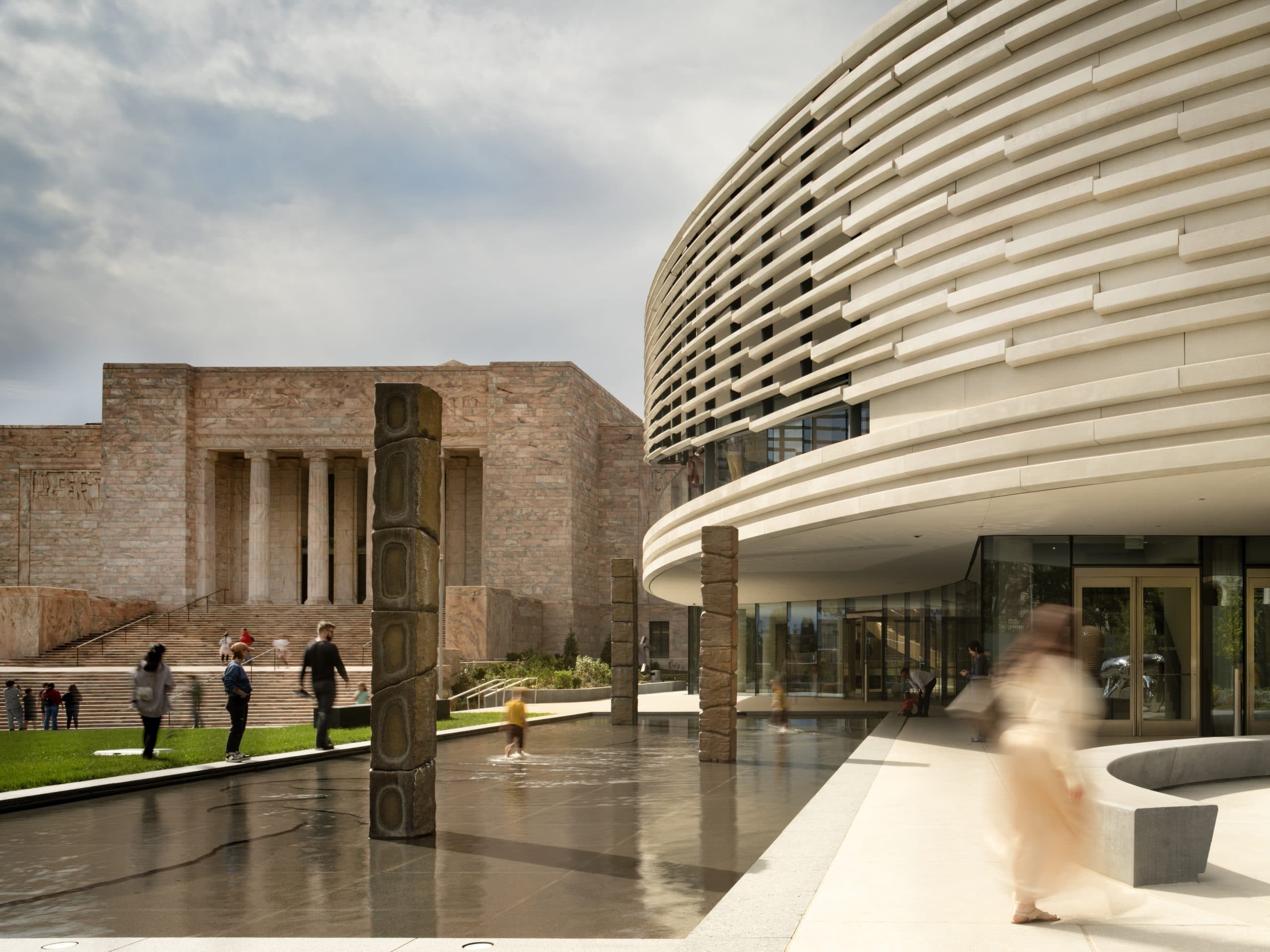 A modern curved building with horizontal lines stands beside a reflecting pool, while people walk near the water. In the background, there is a large stone building with columns.