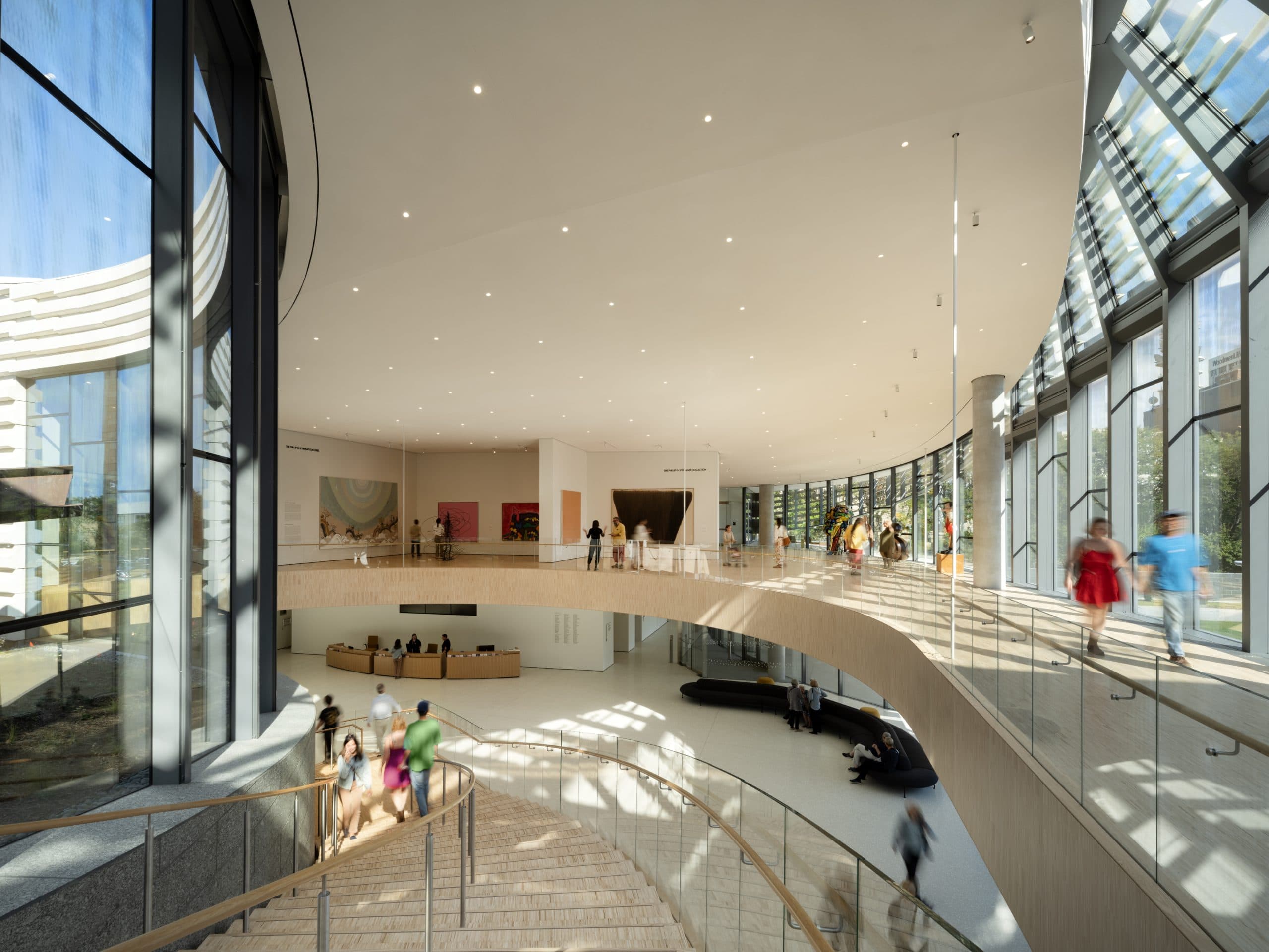 A spacious, modern atrium with large windows, a sweeping staircase, and curved balcony. People are walking and interacting throughout the bright, open space with natural light flooding in.