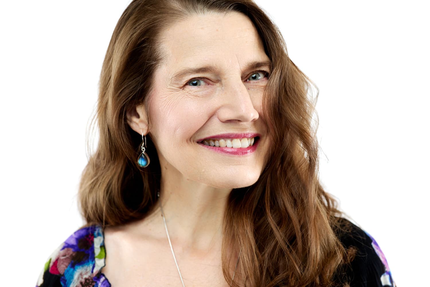 A smiling woman with long brown hair, wearing a colorful top and blue teardrop earrings, poses against a plain white background.