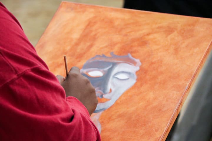 A person in a red shirt paints a portrait of a face with closed eyes on an orange canvas, using a thin brush. The painting appears unfinished, with the face showing subtle gray and brown tones.