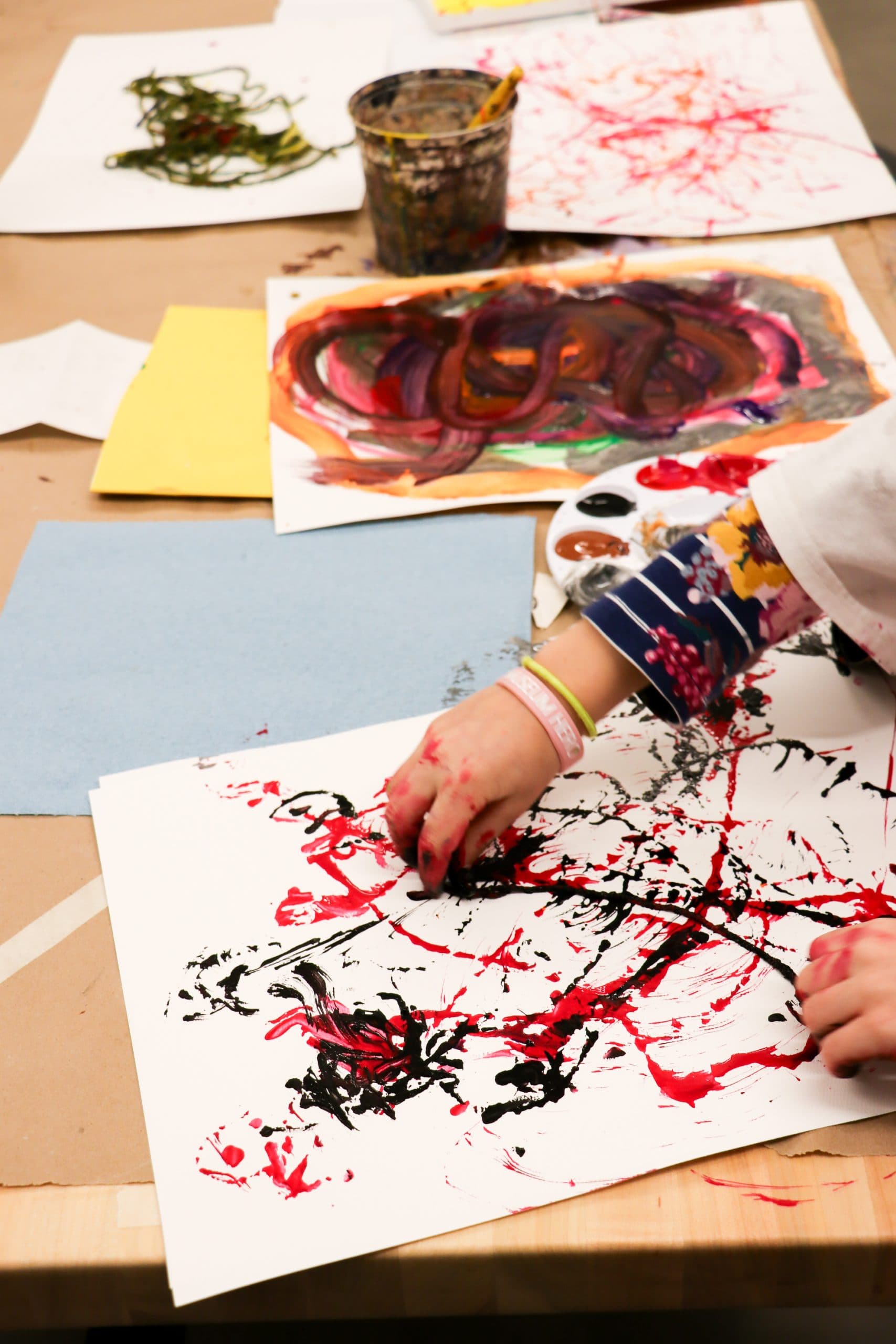 A child’s hands paint on white paper with red and black paint, surrounded by colorful abstract artwork and art supplies on a table. The scene shows a creative art activity in progress.