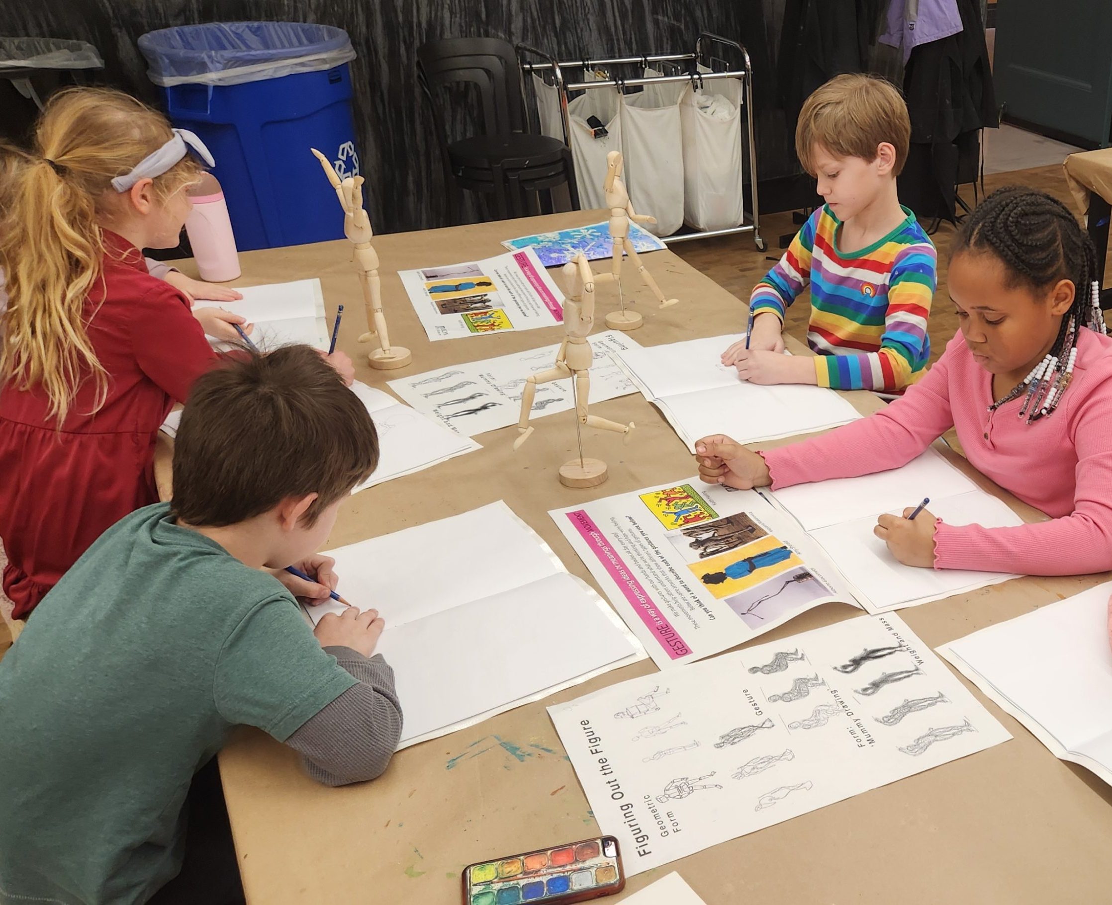 Five children sit around a table drawing on paper, with wooden artist mannequins and drawing examples in front of them. Art supplies and watercolors are visible on the table. The setting appears to be a classroom or art studio.