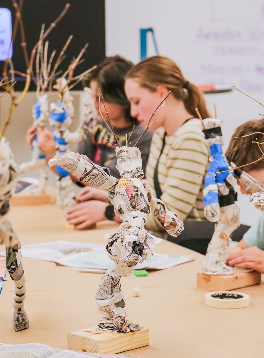 Children sit at a table crafting sculptures made of newspaper and tape on wooden bases. Focus is on a foreground sculpture resembling a dancing figure with outstretched arms and twigs for fingers.
