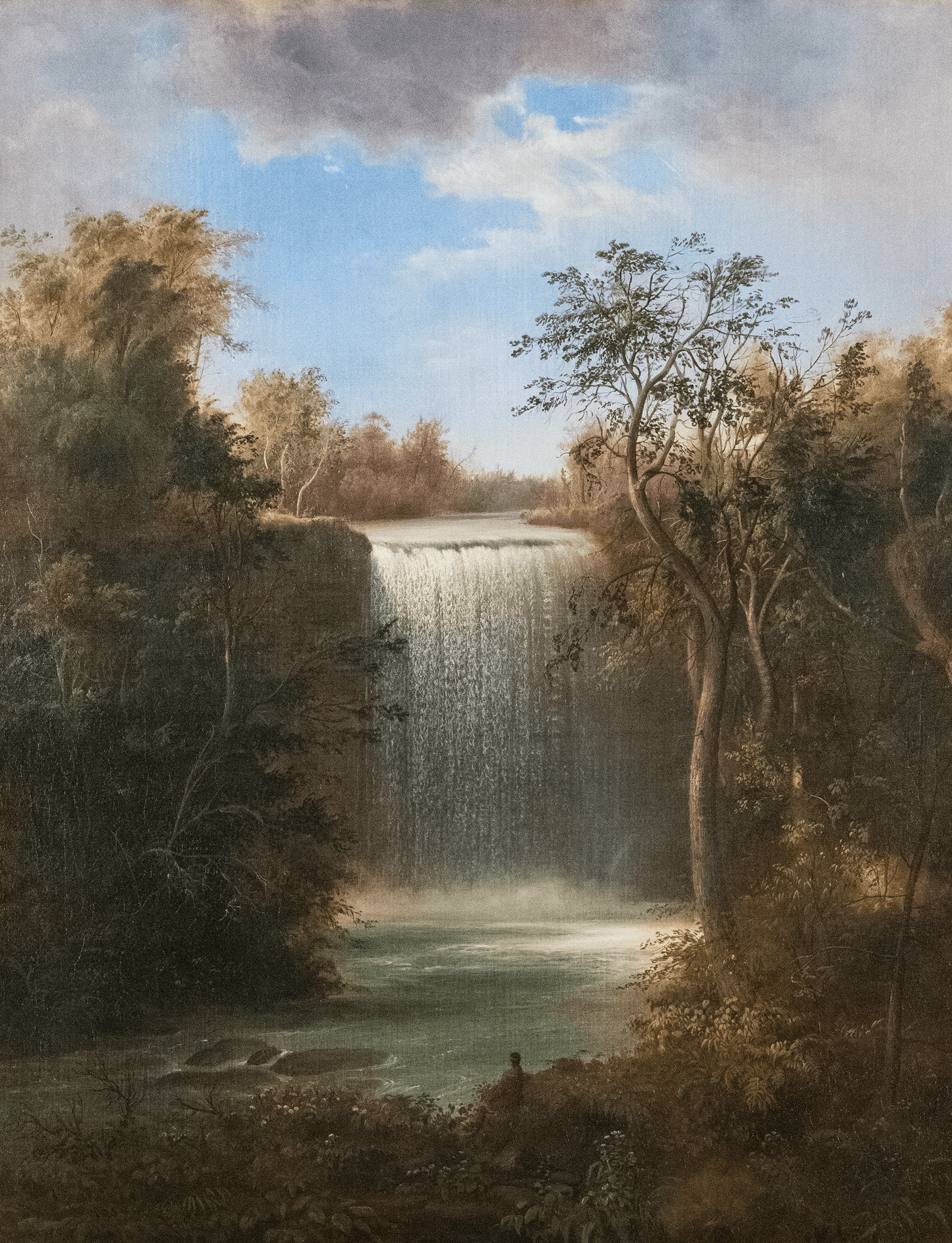 A tall waterfall cascades into a calm pool surrounded by lush, green trees with a partly cloudy sky above. A person stands near the water's edge, admiring the natural scene.