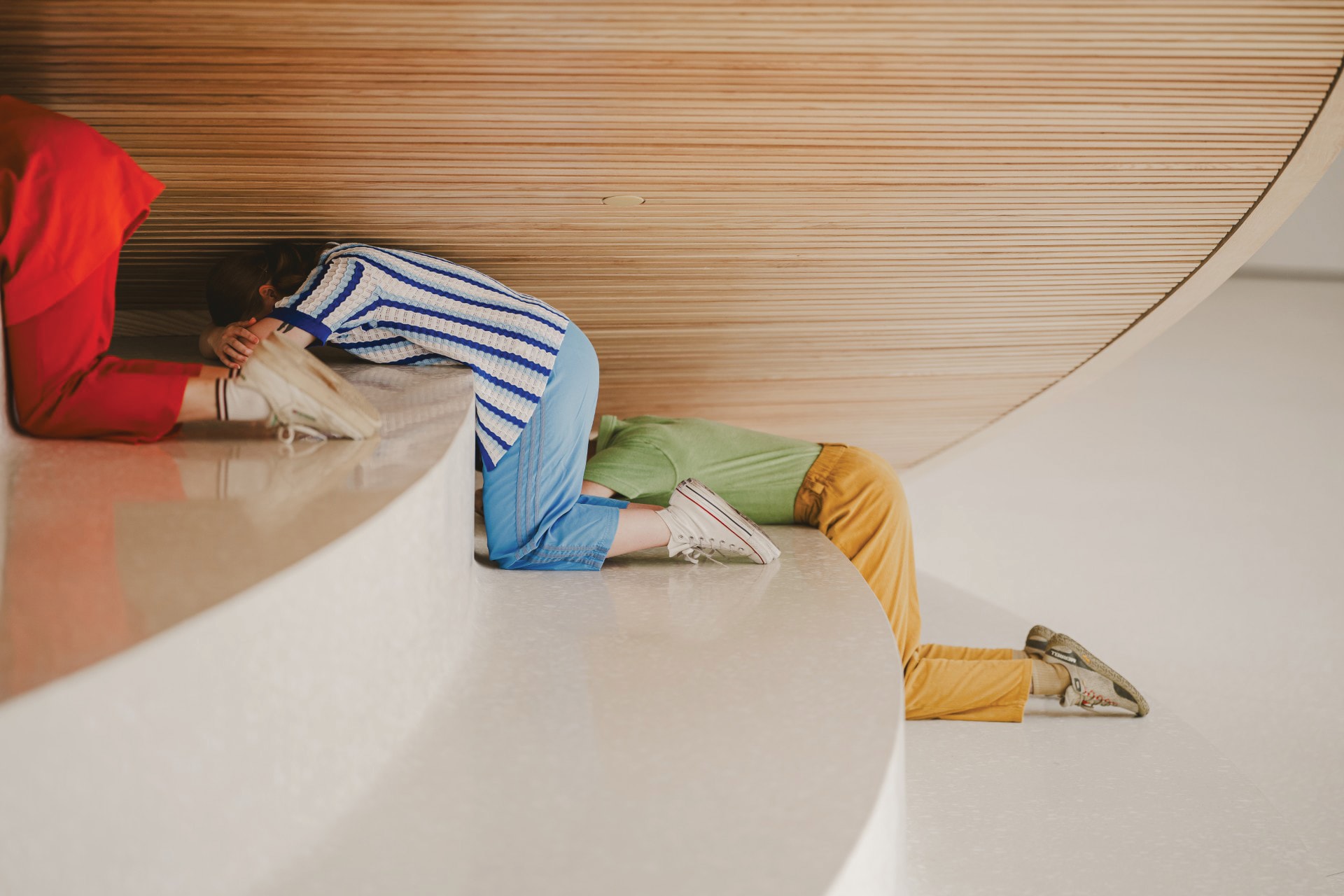 Three people crouch and crawl under a curved, wooden architectural structure, partially hidden. They wear colorful clothing, including a striped shirt, yellow pants, and sneakers, on a smooth, modern floor.