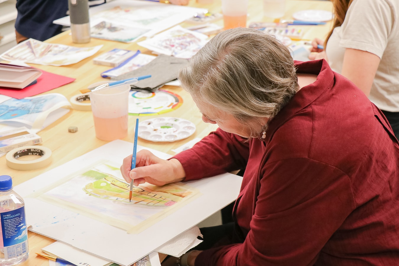 An older woman with gray hair, wearing a red shirt, paints a watercolor picture at a table covered with art supplies and paper, surrounded by other people working on similar projects.