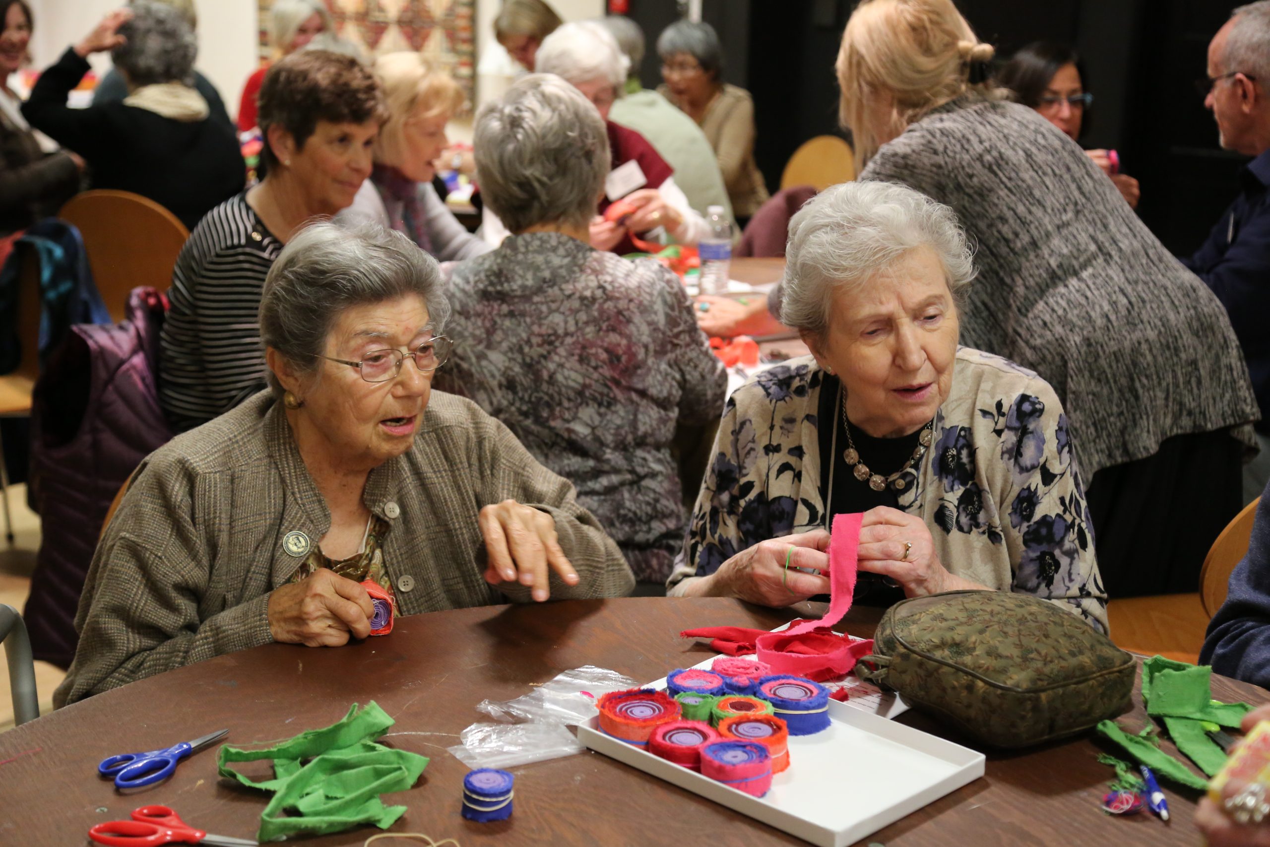 A group of elderly women sit around tables, crafting with colorful felt and fabric. Two women in the foreground are focused on their work, surrounded by handmade felt decorations and craft supplies.