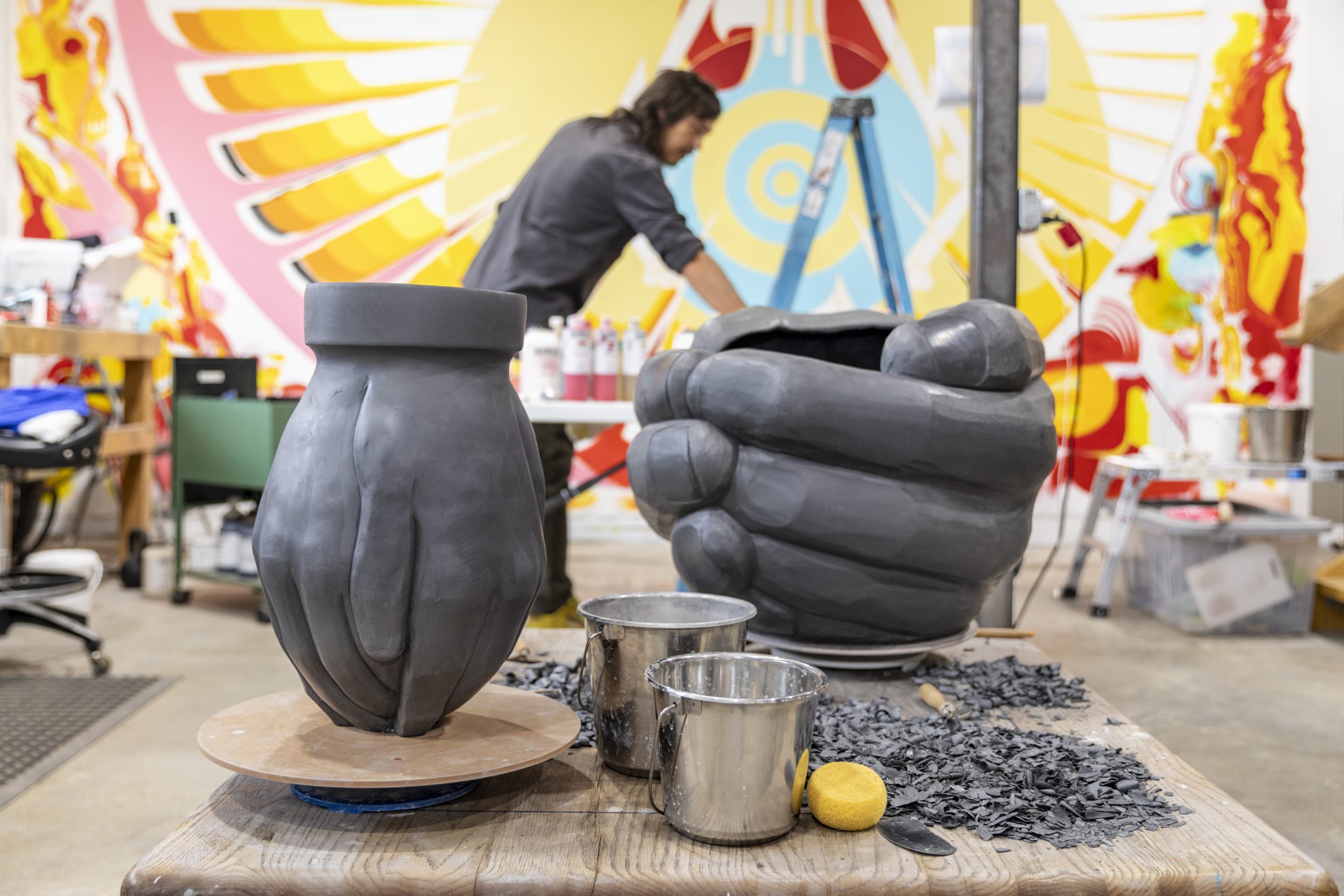 An artist works in a colorful studio with large sculptures of gray hands in the foreground. Buckets, a lemon, and scattered black materials rest on a worktable. A vibrant mural covers the wall behind.