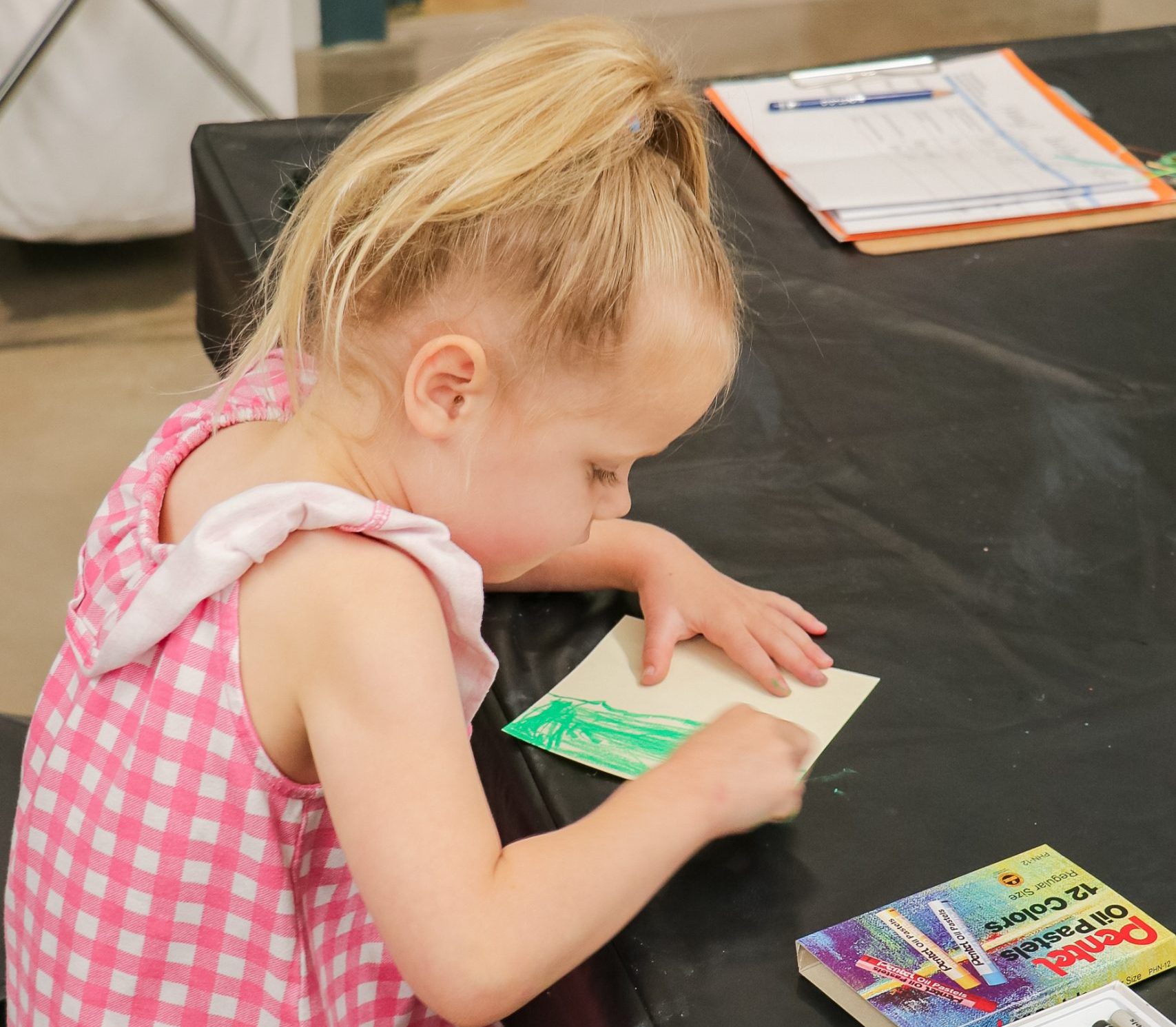 A young girl in a pink checkered dress sits at a table coloring on a piece of paper with oil pastels. Art supplies and papers are spread on the black tablecloth in front of her.