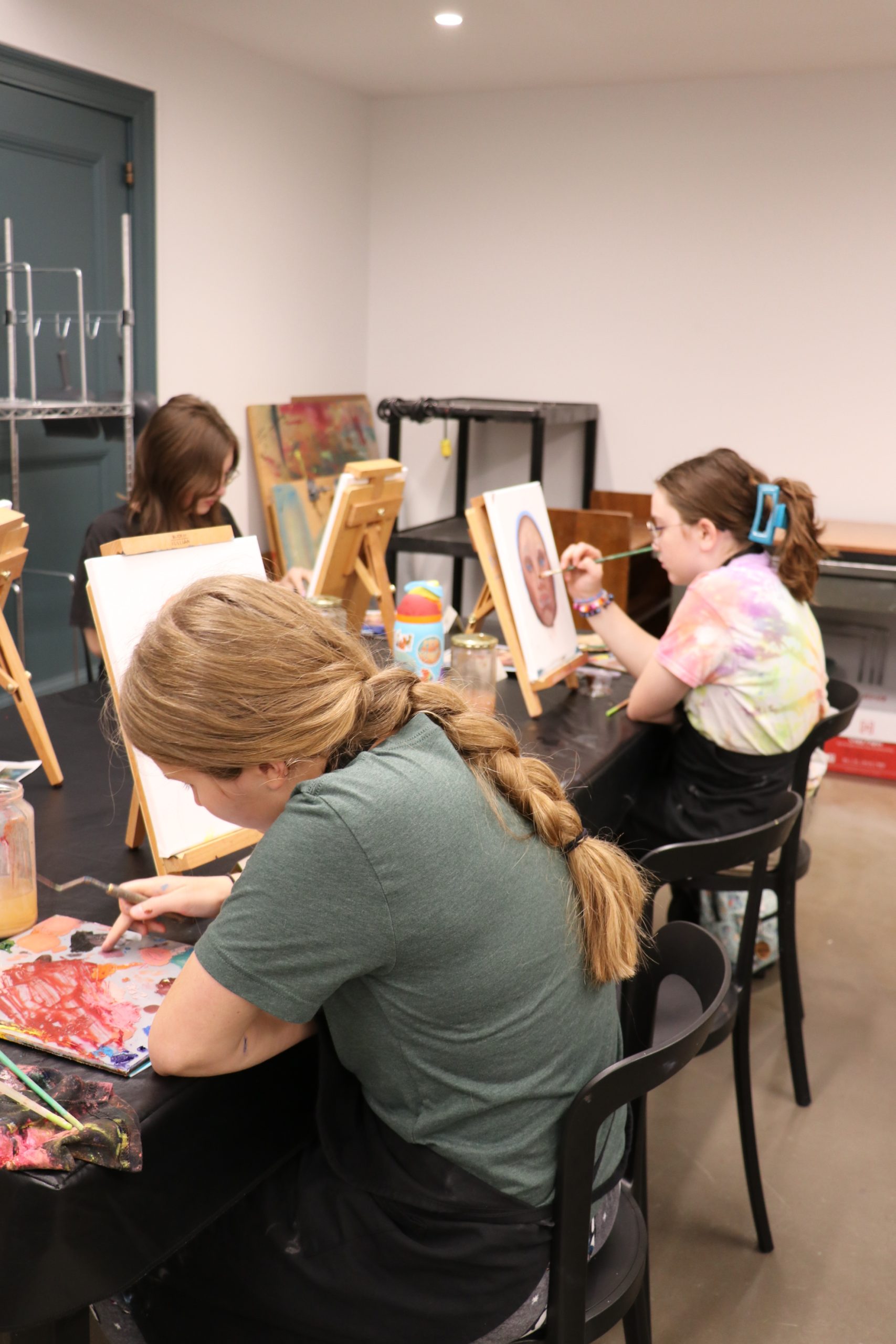 Three people sit at a table painting on canvases positioned on easels. Art supplies and paint palettes are scattered on the table, and one canvas shows a realistic face. The room has a creative, studio-like atmosphere.