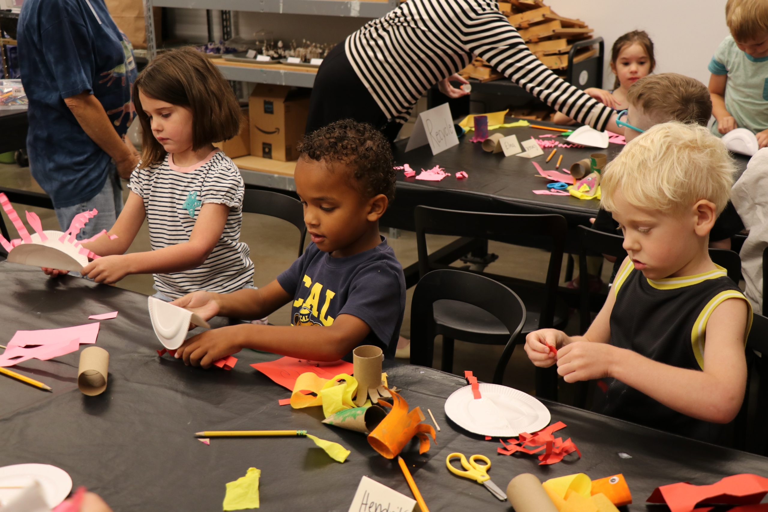 Several young children sit at a table covered with art supplies, such as paper, scissors, and pencils, as they create colorful crafts. An adult assists other children in the background.