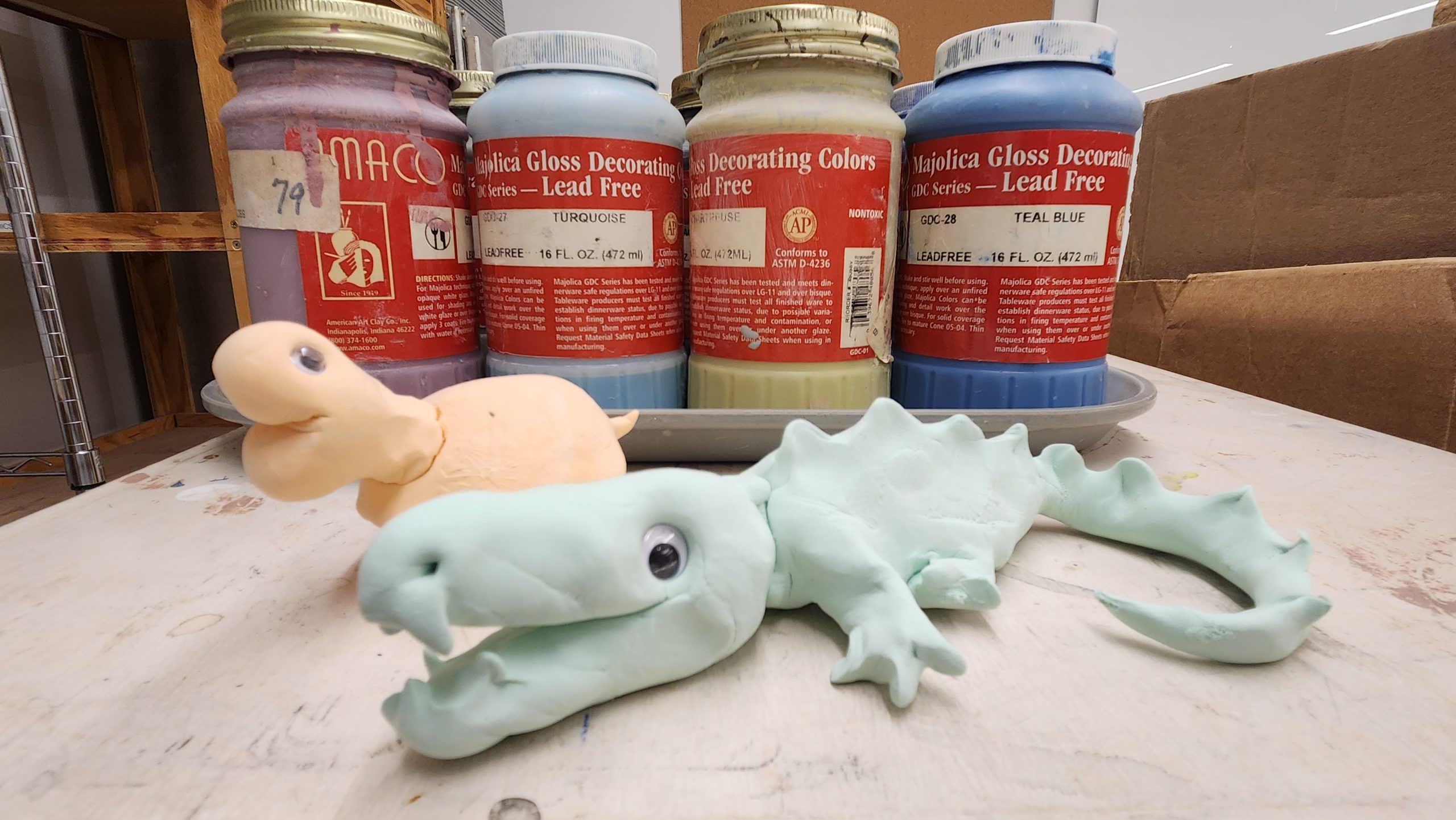Two pastel-colored clay animals, a turtle and a dragon, sit on a table in front of four jars of colorful ceramic glaze labeled "Kalifca Gloss Decorating Colors.