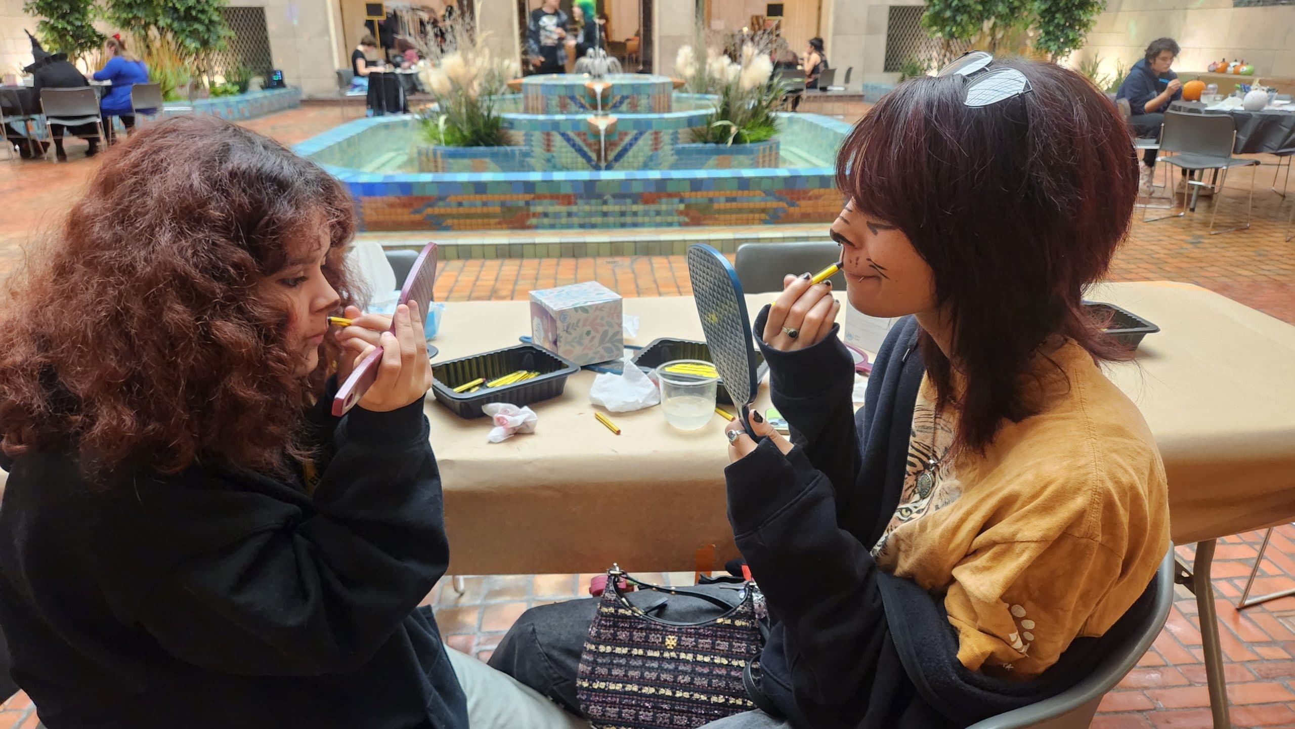 Two people sit at a table in front of a fountain, applying makeup while looking into mirrors. There are food containers on the table, and the setting appears to be an indoor courtyard with plants and people in the background.