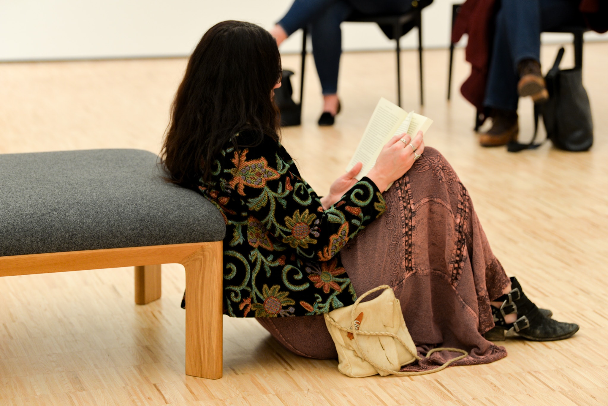 A woman with long dark hair sits on the floor, leaning against a bench, reading a book. She wears a patterned jacket, a long skirt, and black shoes, with a beige bag beside her. Other people are sitting in the background.