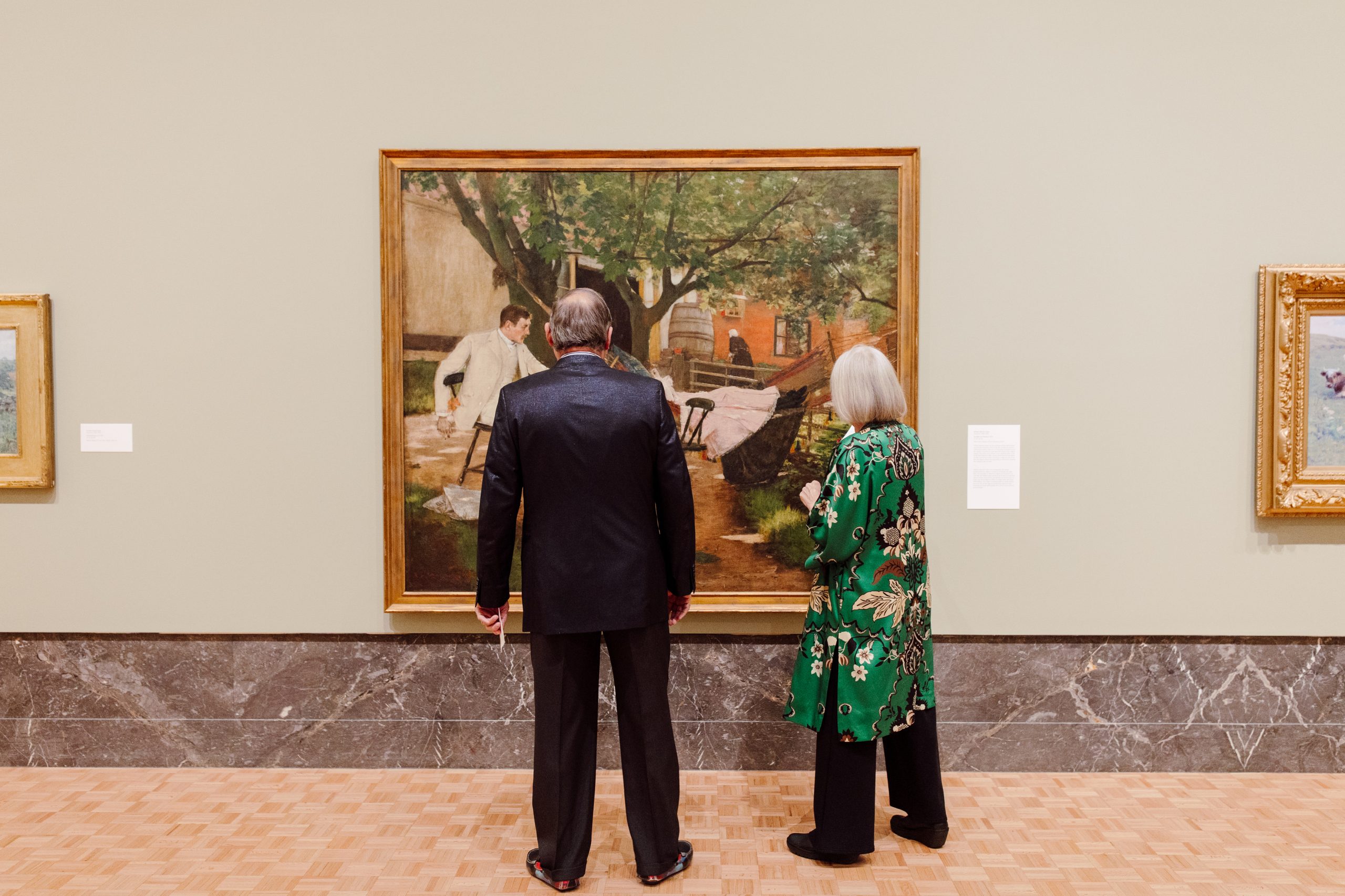 Two people stand side by side closely observing a large framed painting in an art gallery. The painting depicts figures in an outdoor setting with trees. The wall and floor are neutral tones.