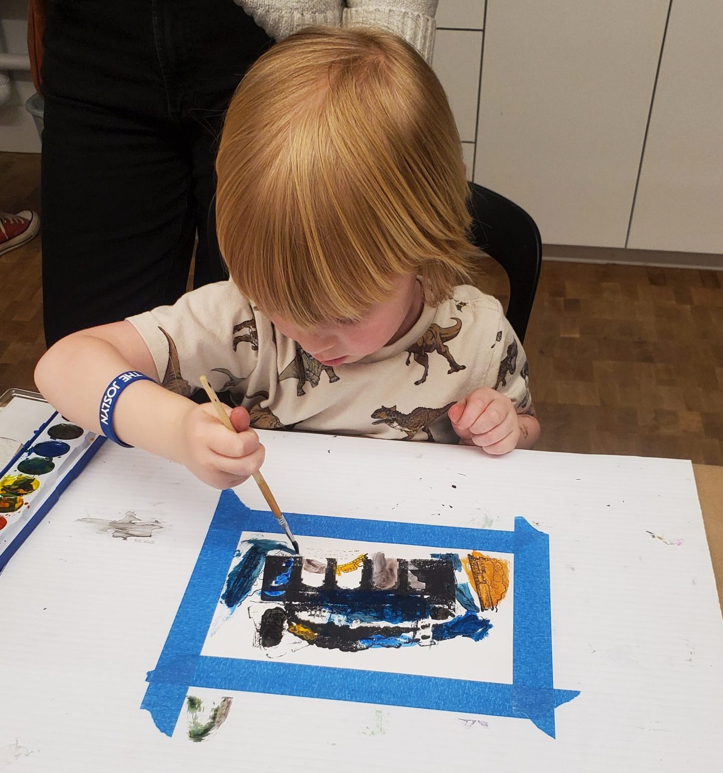 A young child with red hair paints on a piece of paper taped to a table, using watercolors. An adult stands nearby, and art supplies and a flyer are visible on the table.