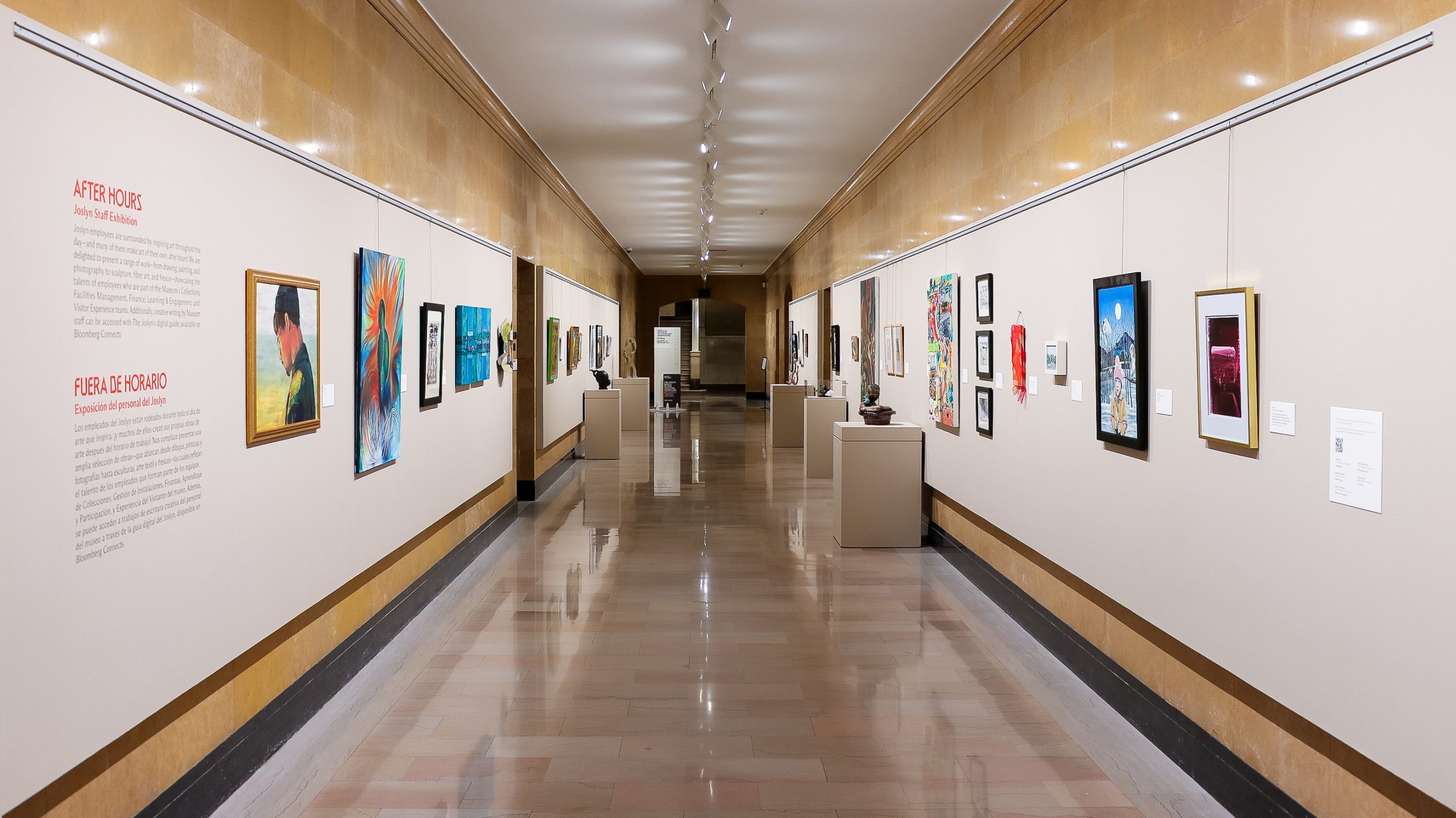 A brightly lit art gallery hallway with shiny floors, displaying various colorful paintings and artworks on both walls; art descriptions and labels are visible alongside the pieces.