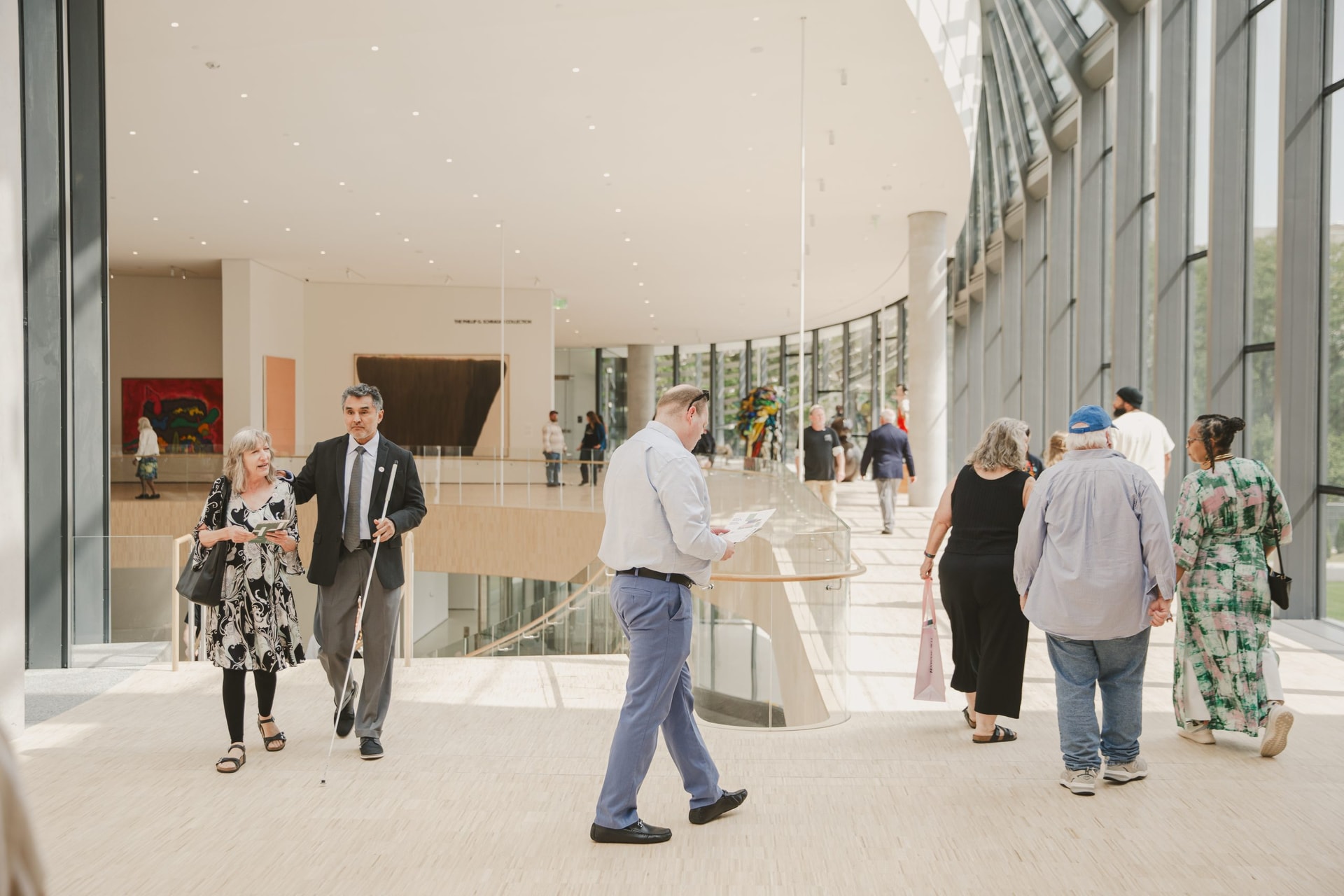 People walk and talk inside a spacious, modern building with large windows, light-colored floors, and natural light streaming in. Some carry papers or bags, and there is artwork visible in the background.