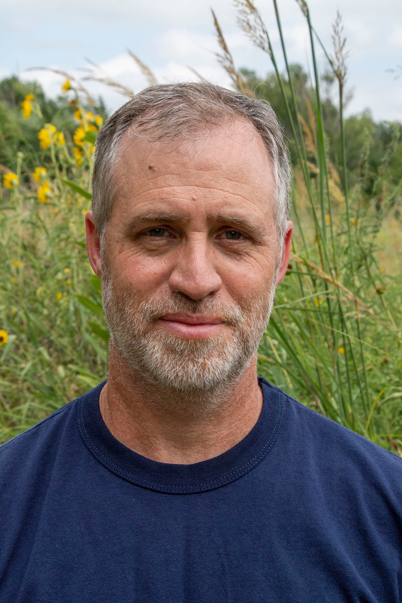 A middle-aged man with short gray hair and a beard, wearing a navy blue shirt, stands outdoors in front of tall grass and yellow flowers on a bright day.