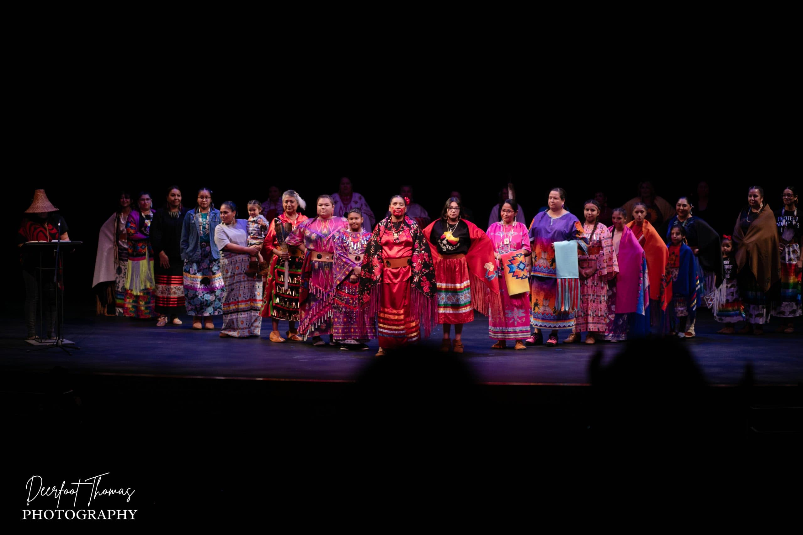 A group of women wearing vibrant, traditional clothing stand in a line on a stage with a dark background, facing the audience under bright stage lights.