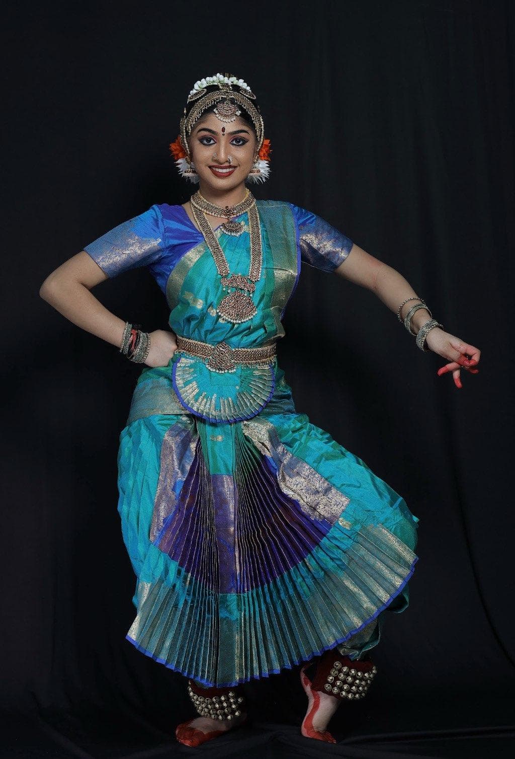 A woman in traditional blue and green Bharatanatyam dance attire poses gracefully against a black background, wearing jewelry, flowers in her hair, and ghungroos on her ankles.