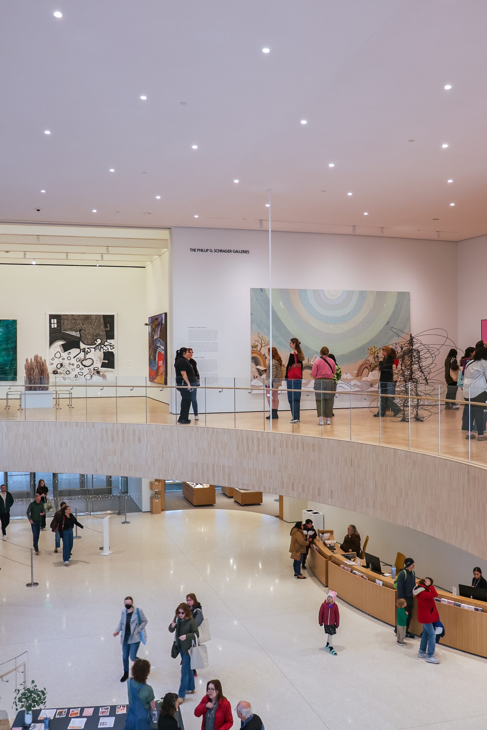 A spacious art gallery with visitors exploring two floors; the lower level has a curved desk and people walking, while the upper level features modern paintings and people observing the artwork.