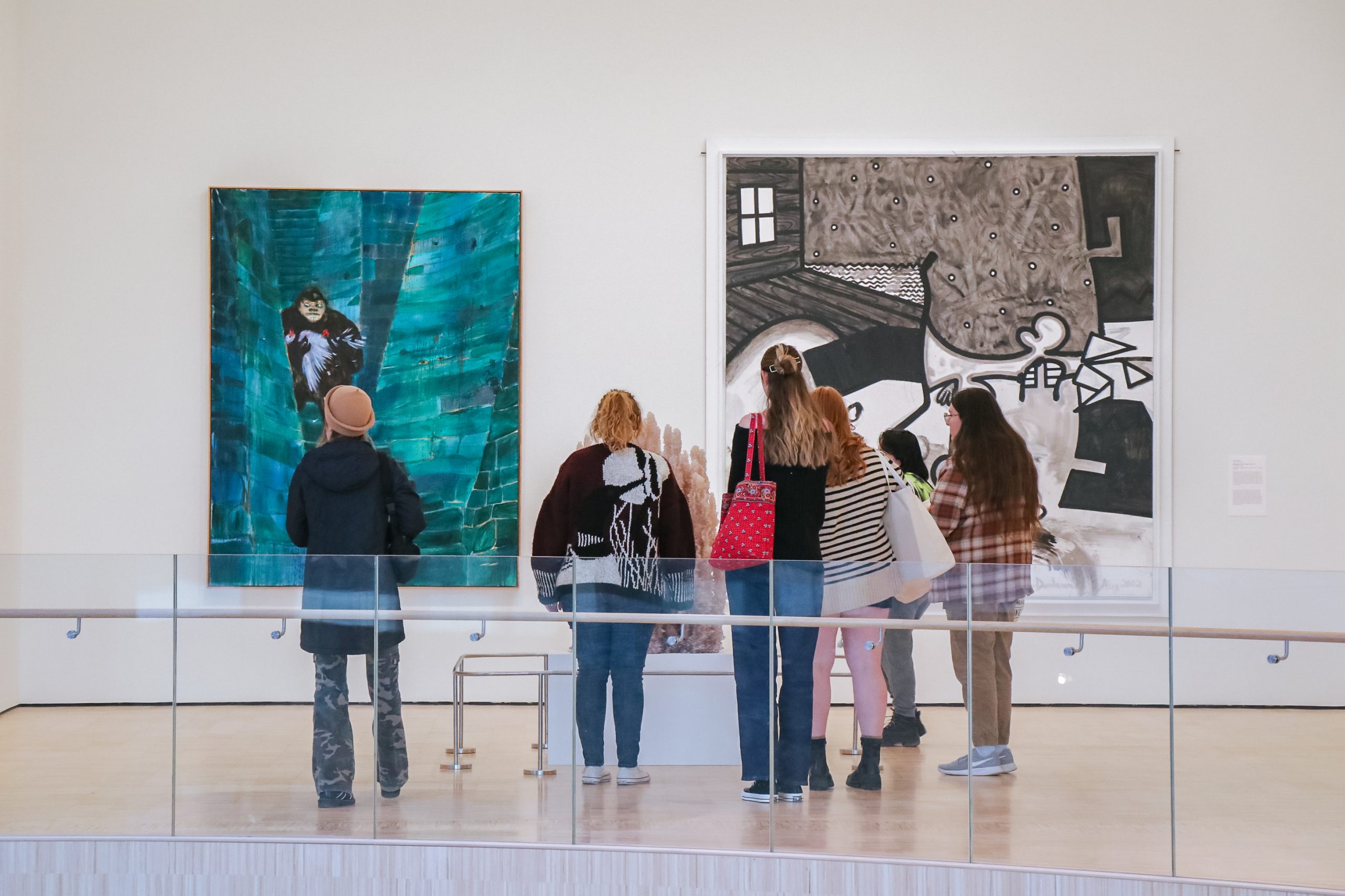 Five people stand in an art gallery, viewing and discussing two large paintings displayed on a white wall, one with predominantly blue tones and the other with black, white, and brown abstract elements.