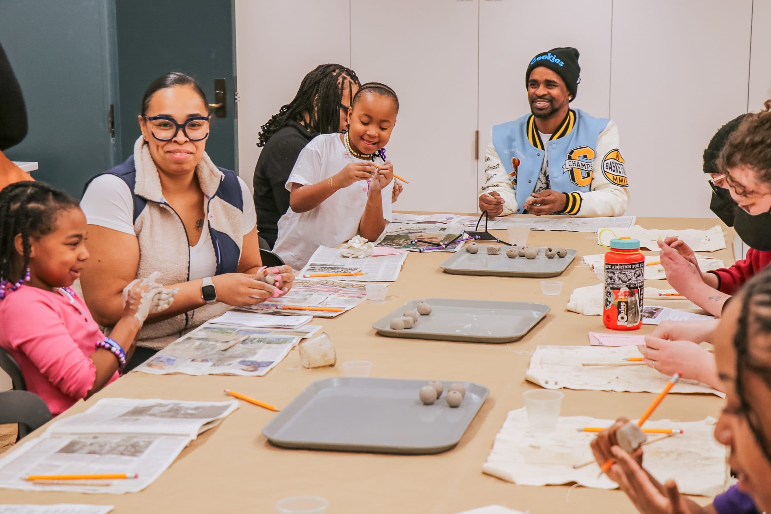 A group of adults and children sit around a table working with clay, smiling and laughing together in a bright, indoor arts and crafts setting. Various clay pieces, pencils, and newspapers are on the table.