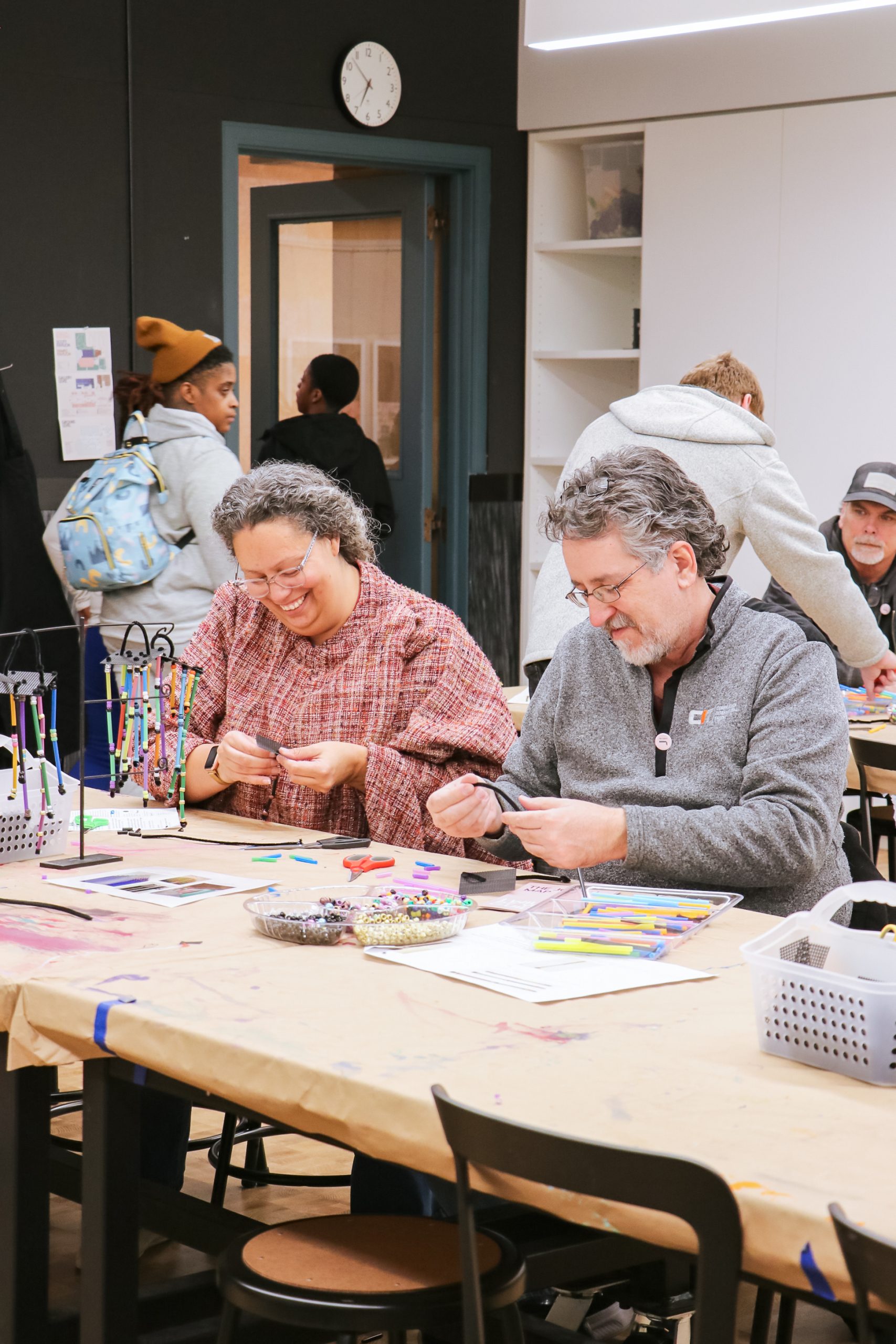 Two adults sit at a table crafting beaded bracelets, smiling and focused. Supplies like beads, string, and tools are scattered on the table. Other people are visible in the background inside a creative, workshop-like room.