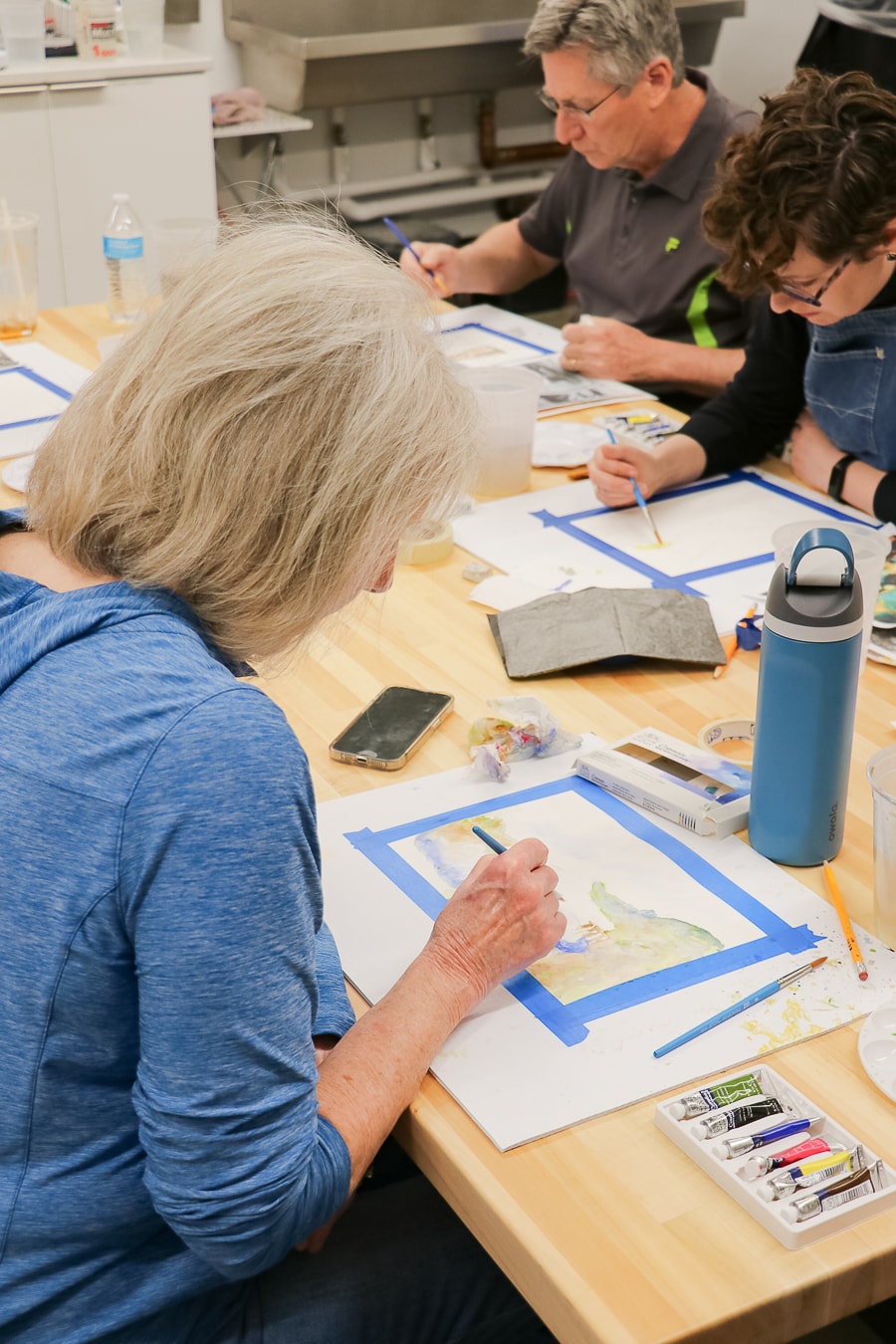 Three people sit at a wooden table, painting with watercolors on paper taped down. Art supplies, brushes, and water bottles are scattered on the table. The focus is on the woman in the blue shirt painting in the foreground.