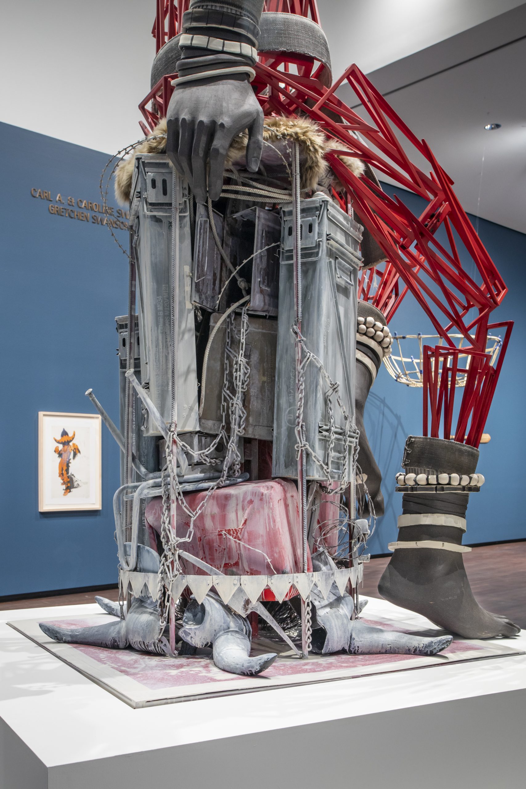 A large, mixed-media sculpture features oversized hands, red geometric rods, and architectural elements resembling metal fencing and columns. The base is surrounded by ropes and rocks. A framed artwork hangs on a blue wall behind it.
