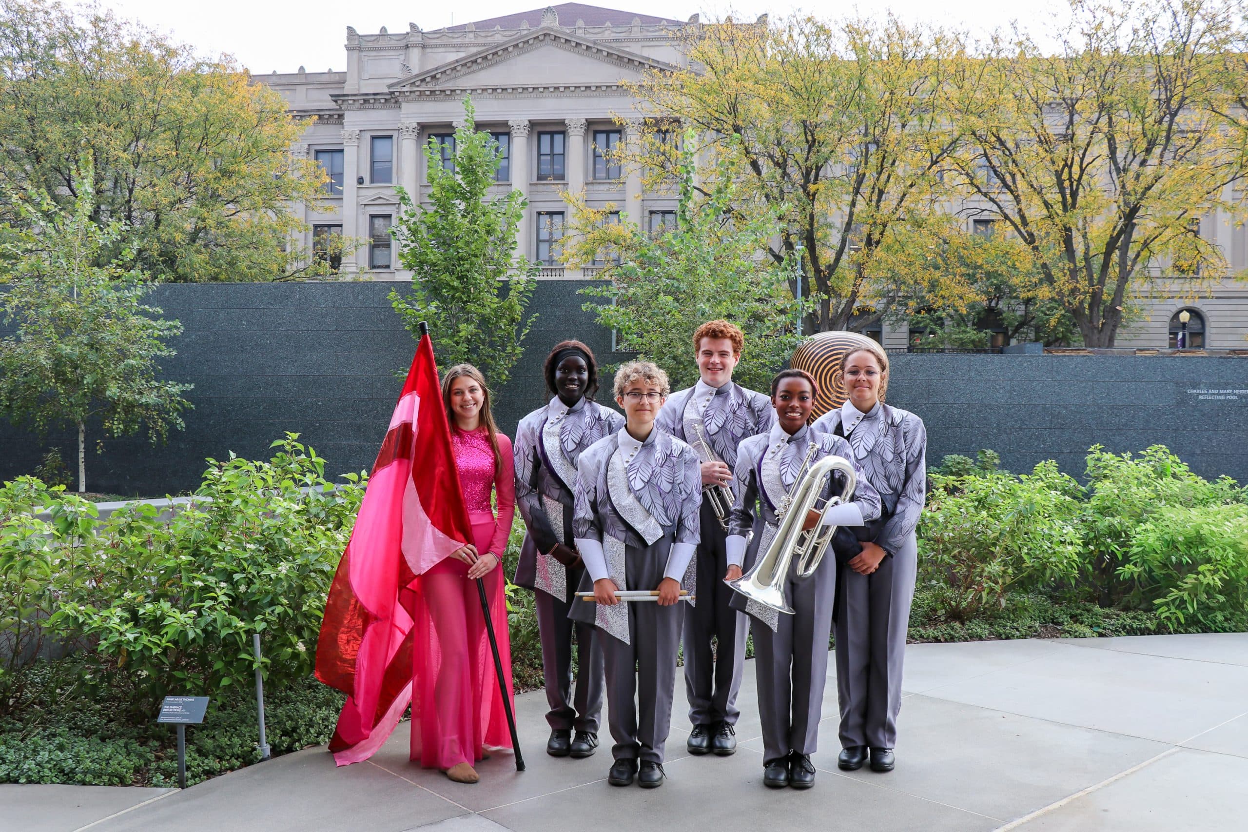 Six students in matching silver marching band uniforms and one in a pink and red flag outfit stand smiling outside in front of a leafy garden, with a large historic building in the background.