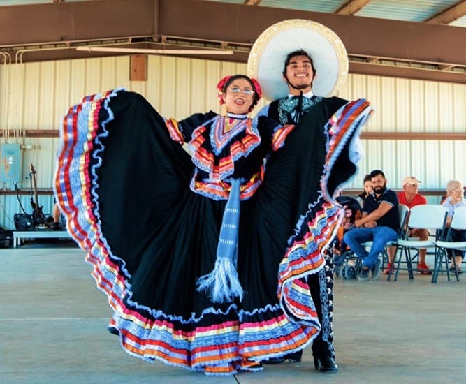 A smiling couple in traditional Mexican folkloric attire poses indoors, with the woman holding out her colorful, ruffled dress and the man wearing a charro suit and sombrero. Seated people watch in the background.