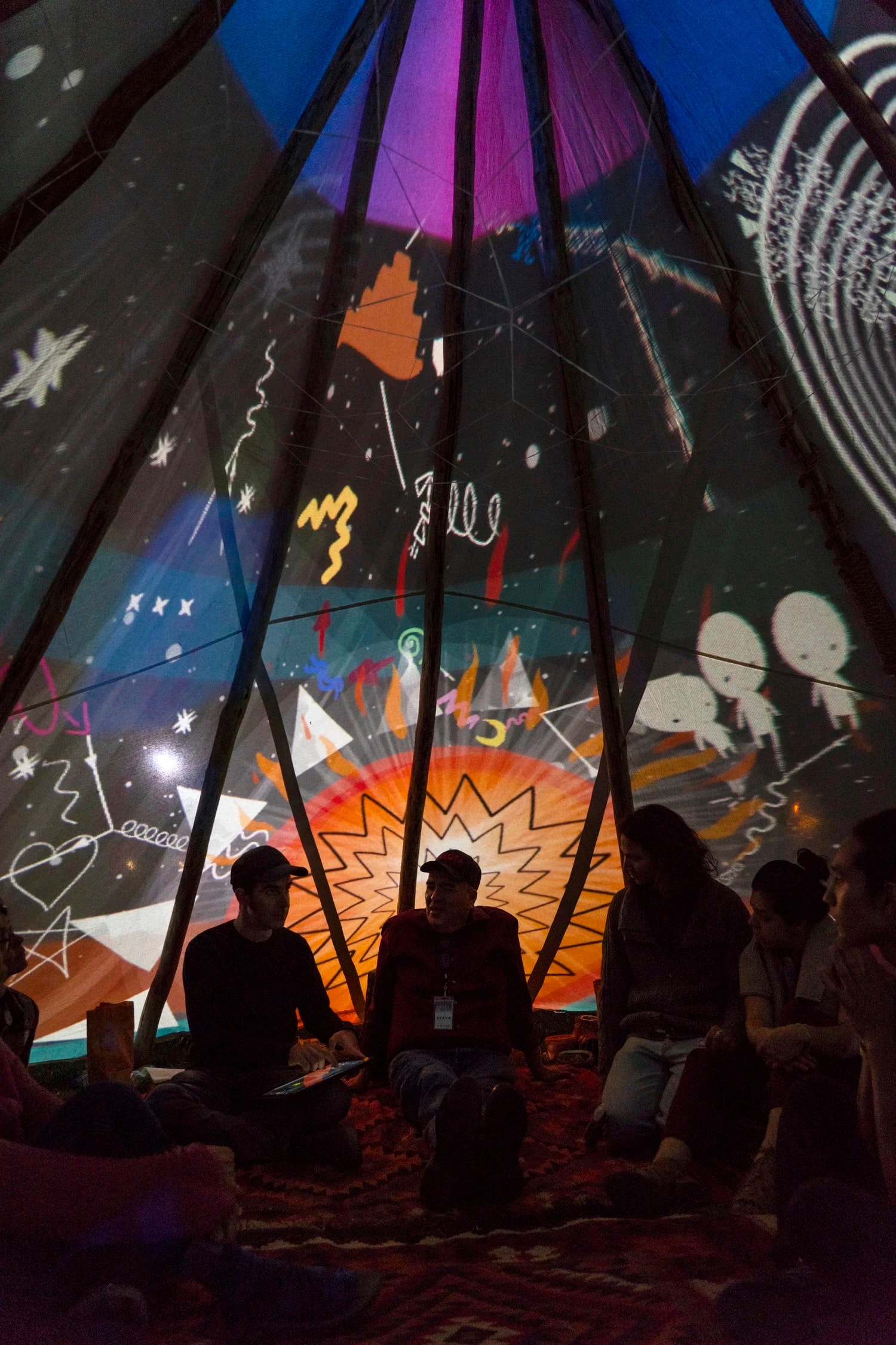 Several people sit inside a tent with colorful, abstract light projections on the fabric above them, including stars, lines, and cartoon-like drawings, creating a vibrant and creative atmosphere.
