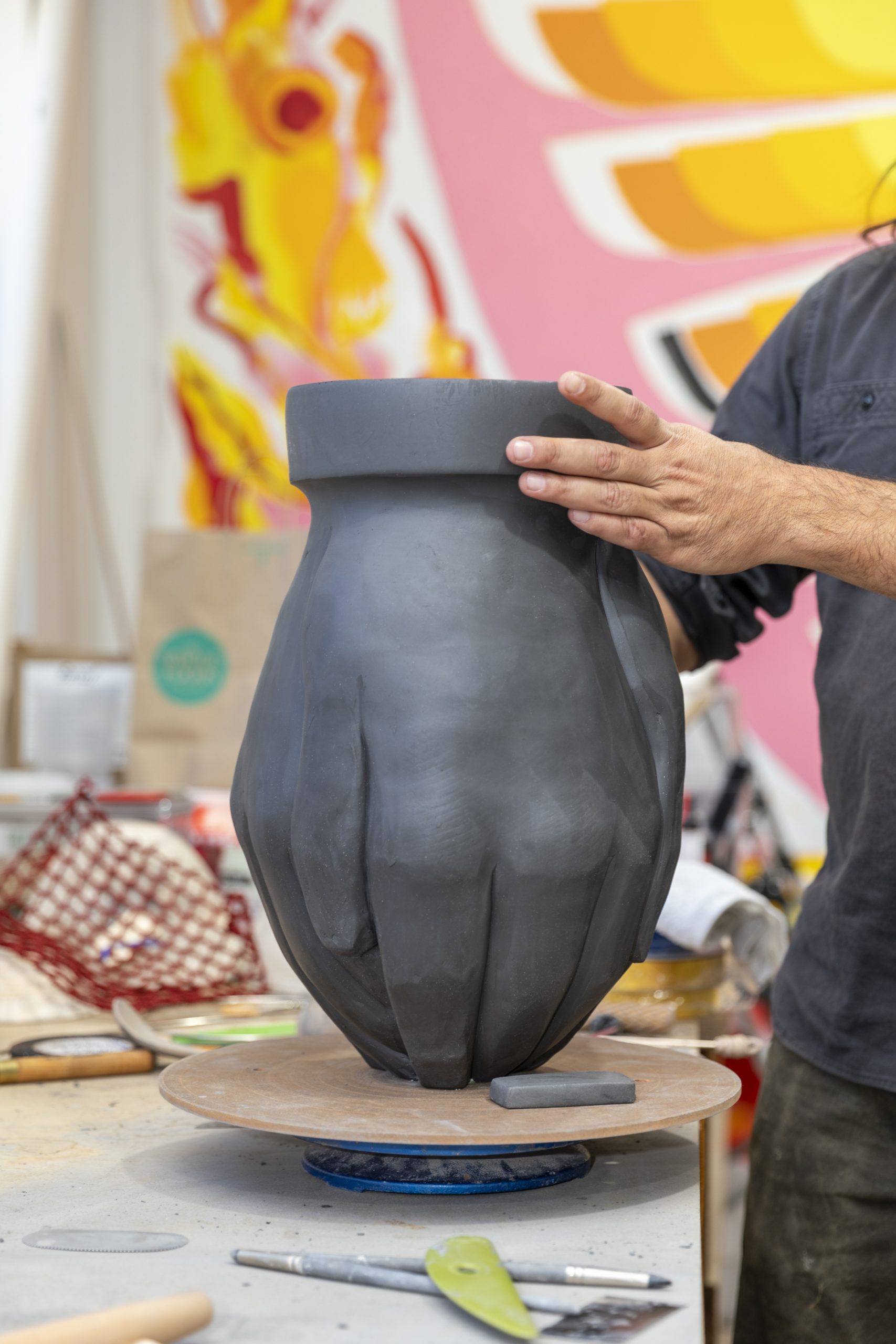 A person shapes a large ceramic sculpture resembling a closed hand in a lively studio, with art supplies and colorful abstract artwork visible in the background.