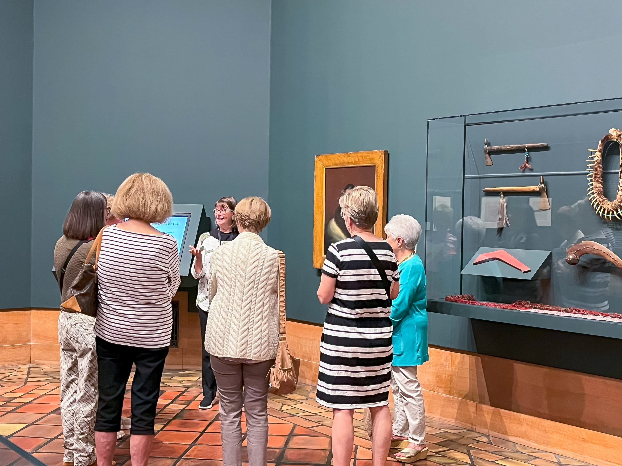 A group of women stand in an art museum, listening to a guide. They face a display case with cultural artifacts and a framed painting on the wall. The floor is patterned, and the walls are painted teal.
